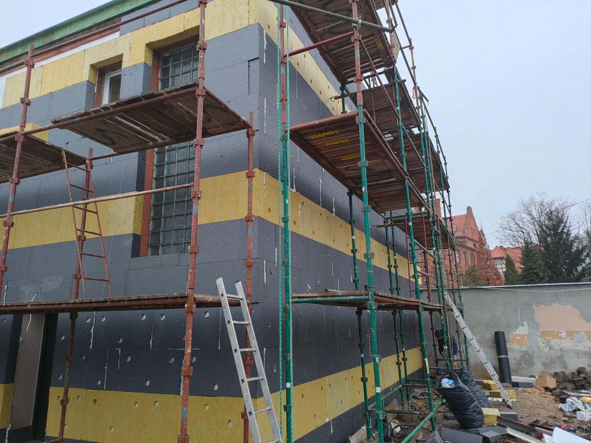 Budynek w trakcie ocieplania: szare i żółte płyty izolacyjne ułożone w pasy, rusztowanie wokół, drabina oparta o ścianę, widoczne okna z luksferami.