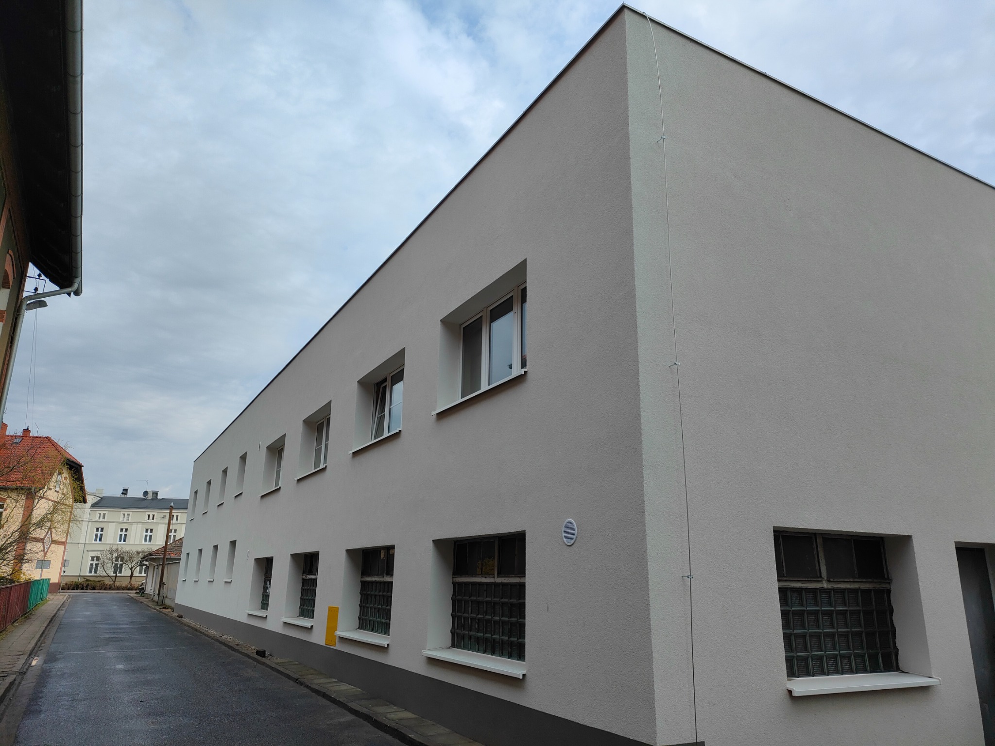 Odnowiony budynek z szarą elewacją, widoczne okna ze szprosami i kratami, tynk strukturalny, ulica po deszczu.