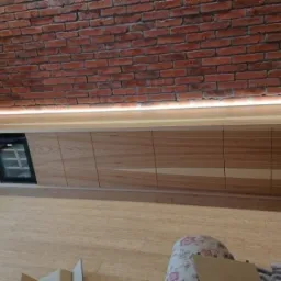 Długa, drewniana zabudowa meblowa pod skosem ściany z czerwonej cegły, z podświetleniem LED, wbudowaną chłodziarką do wina i szafkami.