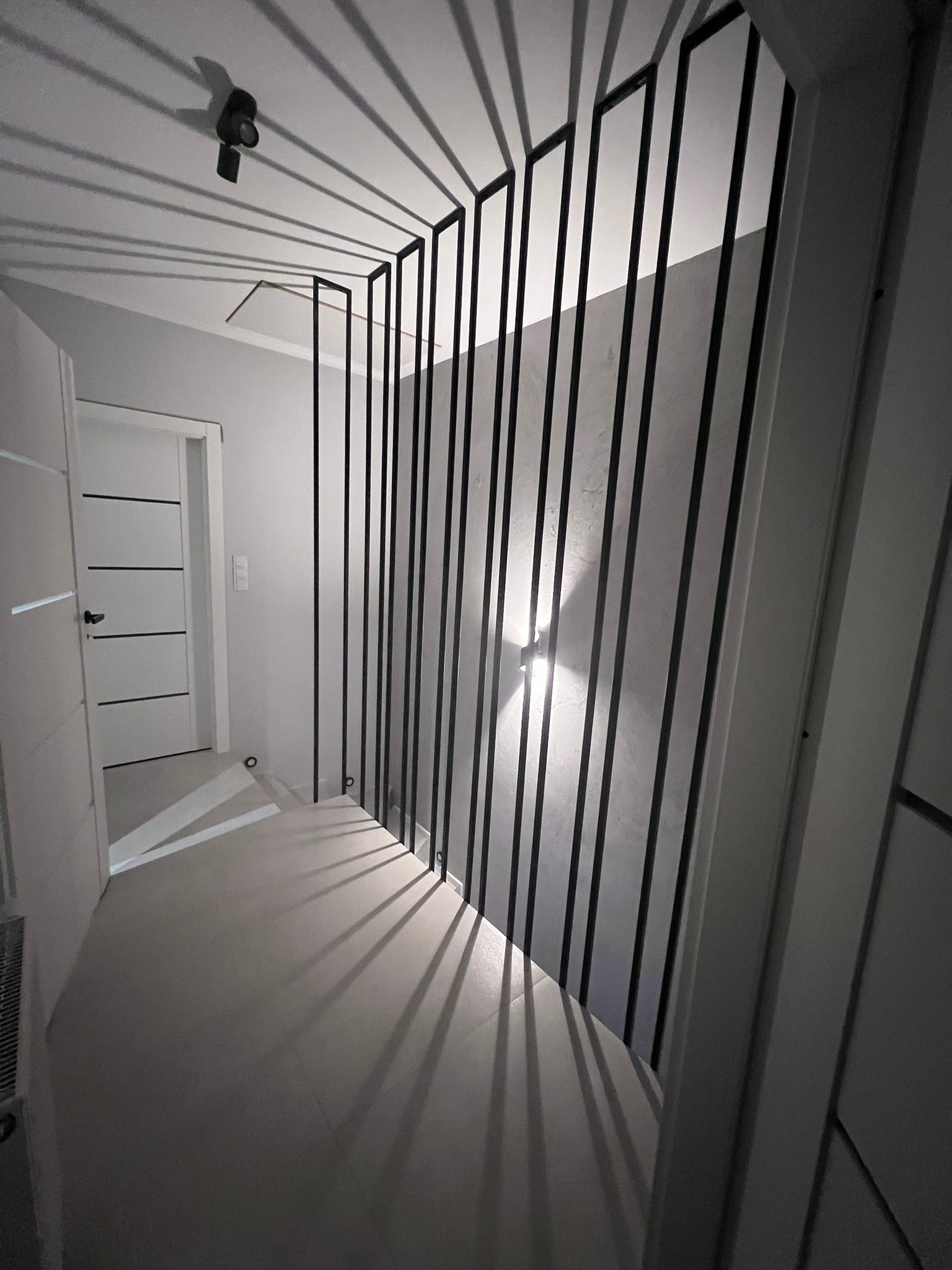 Nowoczesna balustrada schodowa z pionowych, czarnych prętów, tworząca geometryczne cienie na ścianie i podłodze, oświetlona punktowym światłem sufitowym i kinkietem ściennym, widok na białe drzwi...