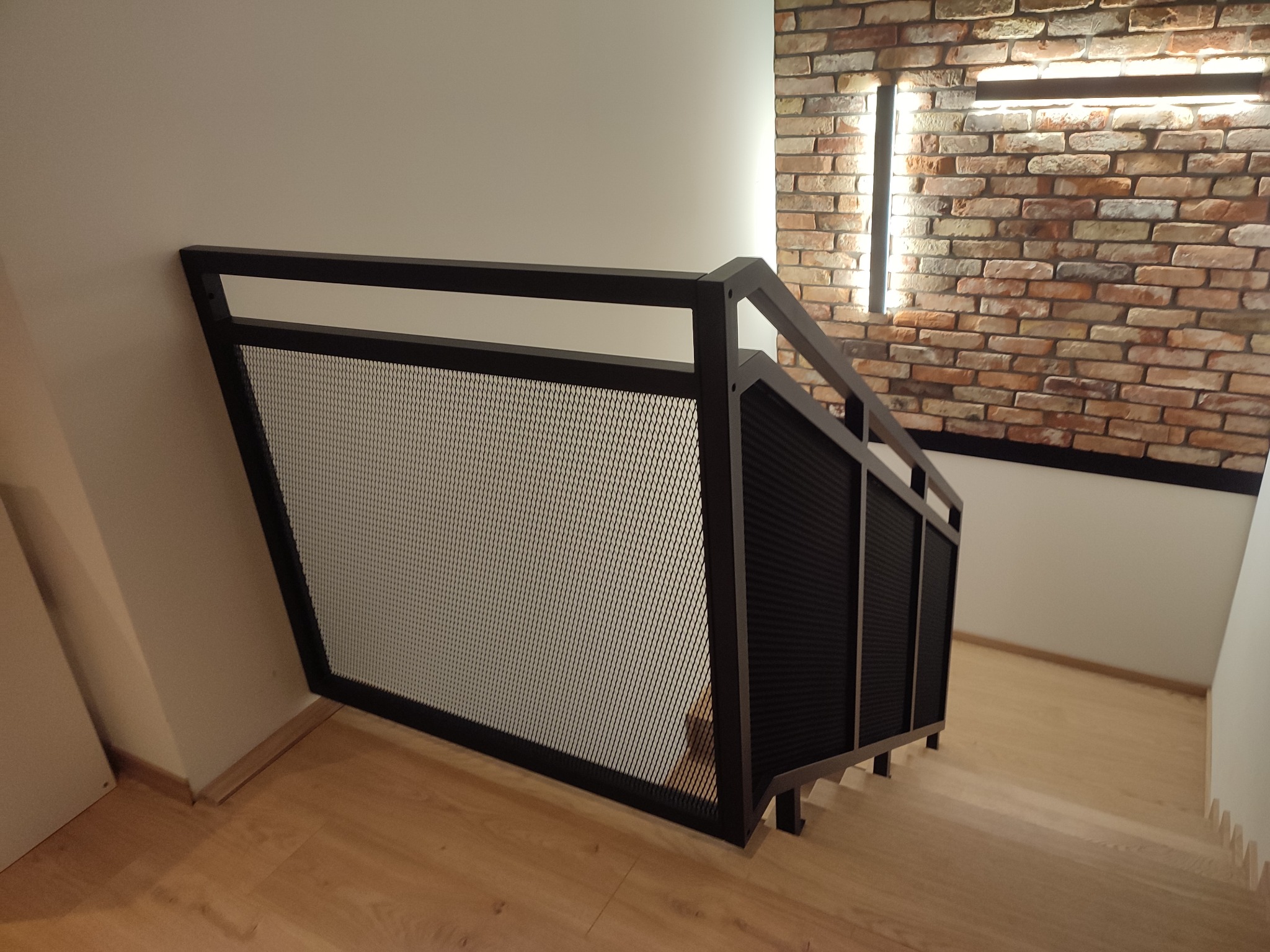 Nowoczesne schody z czarną metalową balustradą z siatkowym wypełnieniem, oświetlenie LED w ścianie z cegły.
