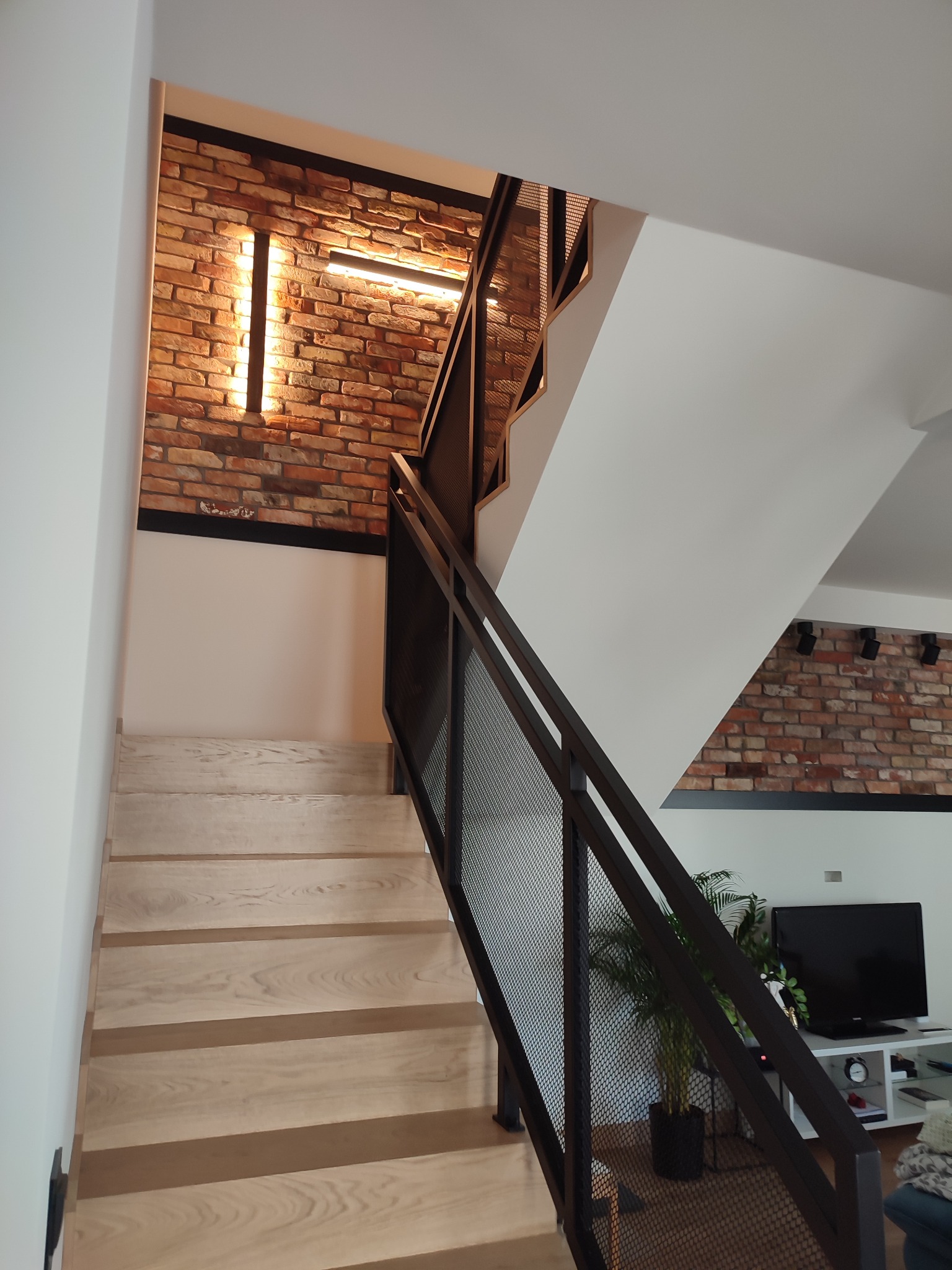Nowoczesne schody drewniane z metalową ażurową balustradą, oświetlenie LED na ceglanej ścianie w tle, widok z dołu.
