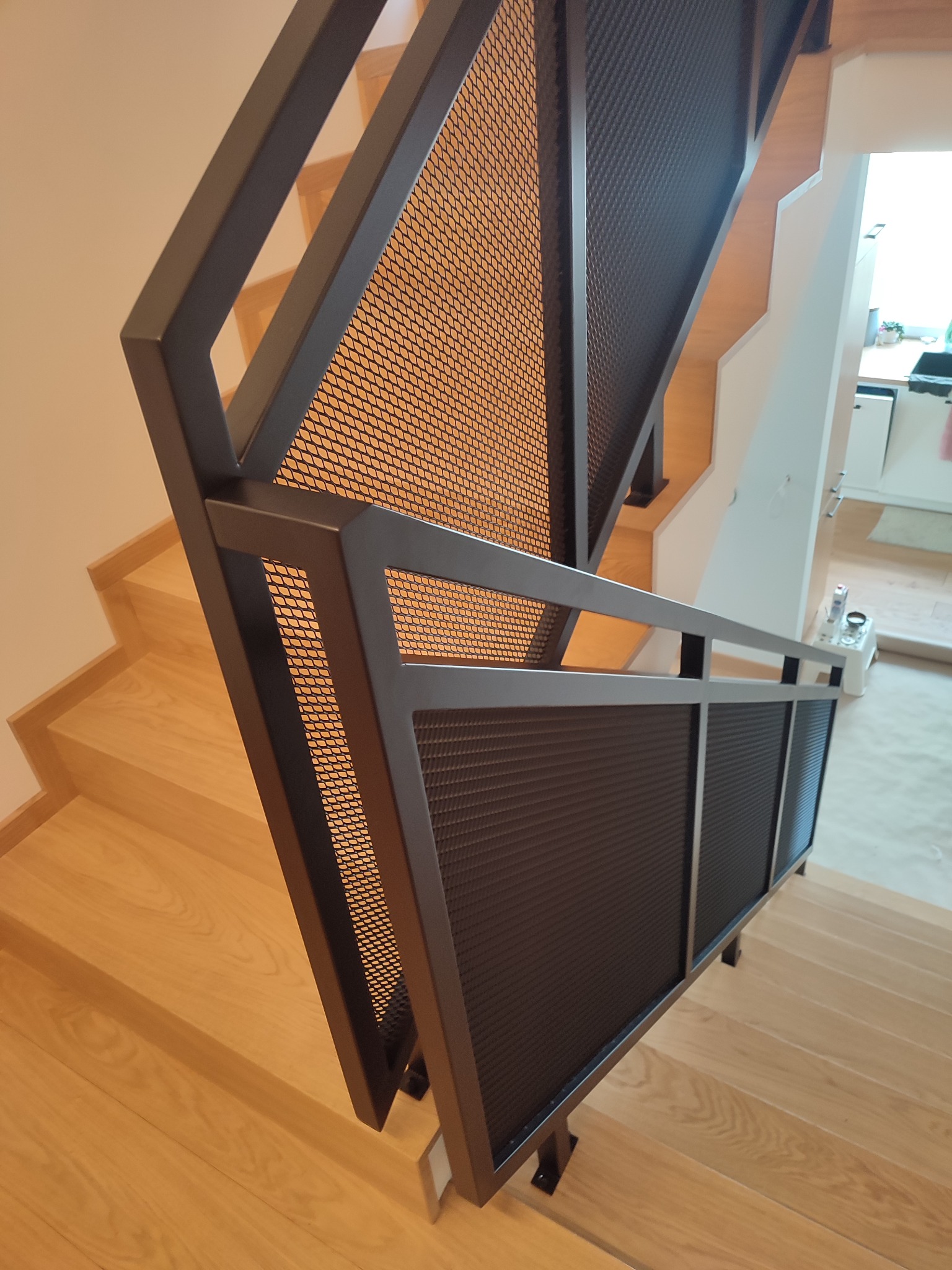 Nowoczesne schody wewnętrzne z drewnianymi stopniami i czarną metalową balustradą wypełnioną siatką, widok z góry na dół.