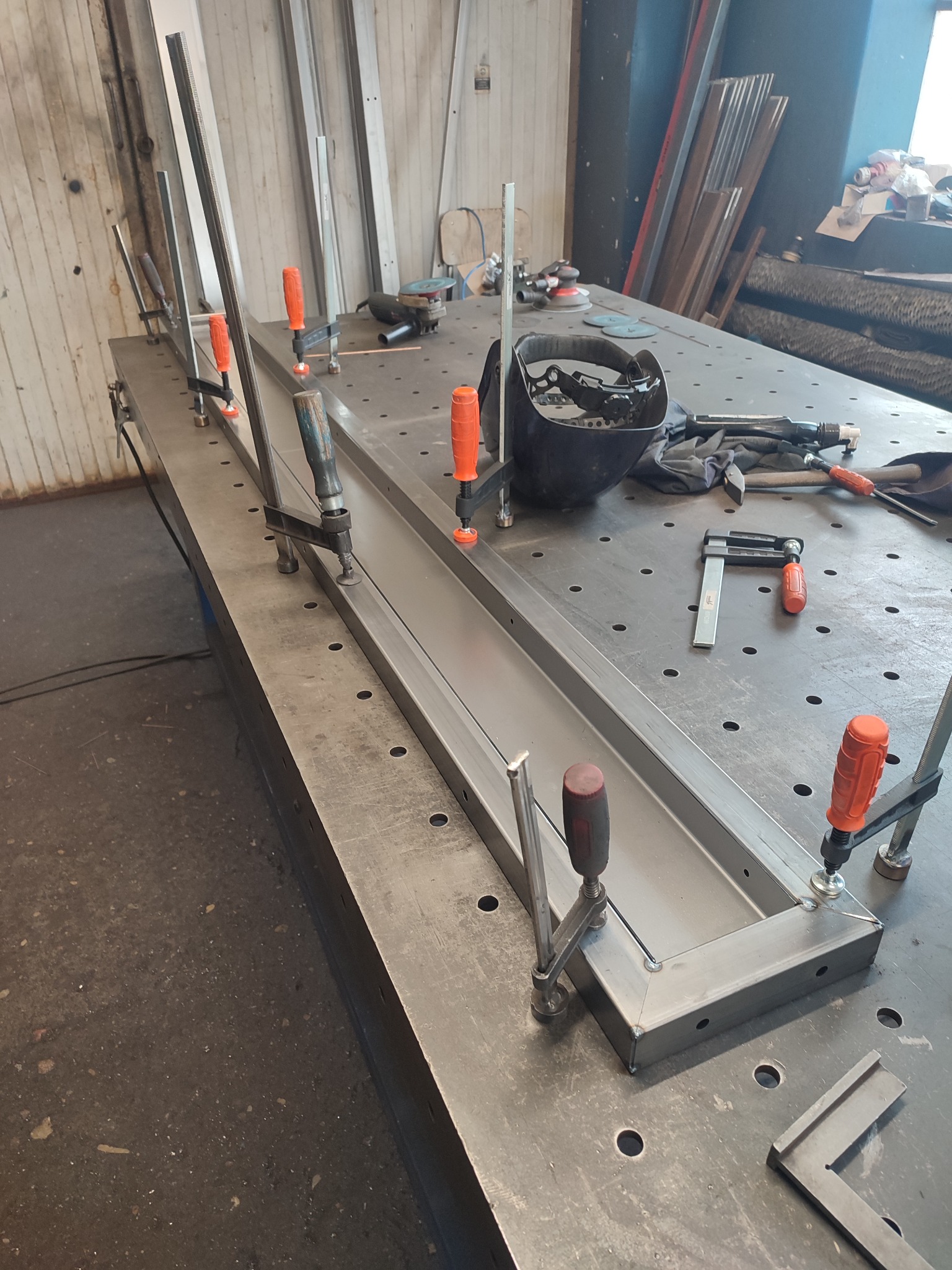 Metalowa rama spawana na stole spawalniczym, zabezpieczona ściskami, z widocznymi narzędziami i maską spawalniczą.