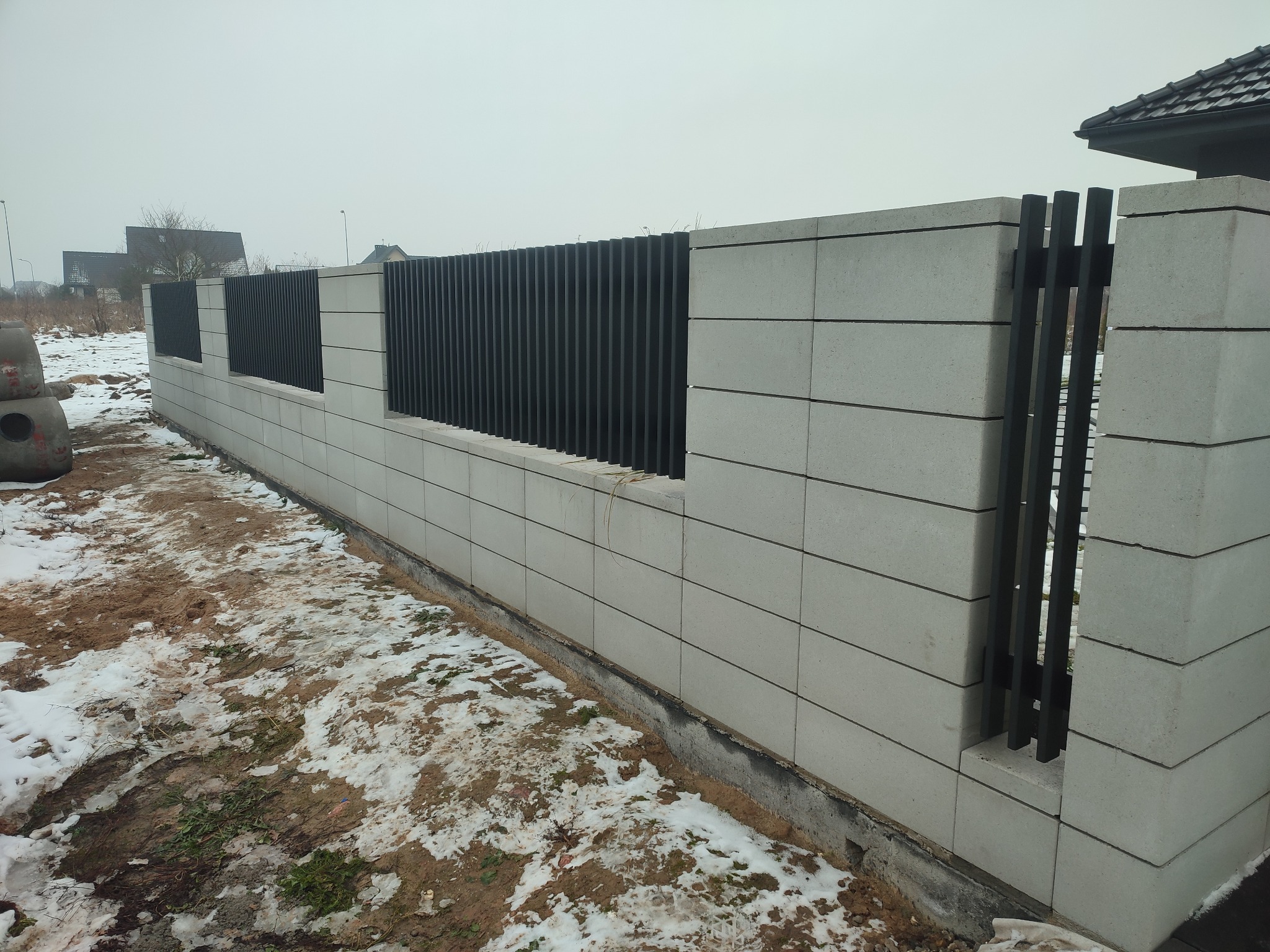 Nowoczesne ogrodzenie z szarego betonu architektonicznego z czarnymi, pionowymi przęsłami, widoczne ślady śniegu na ziemi.