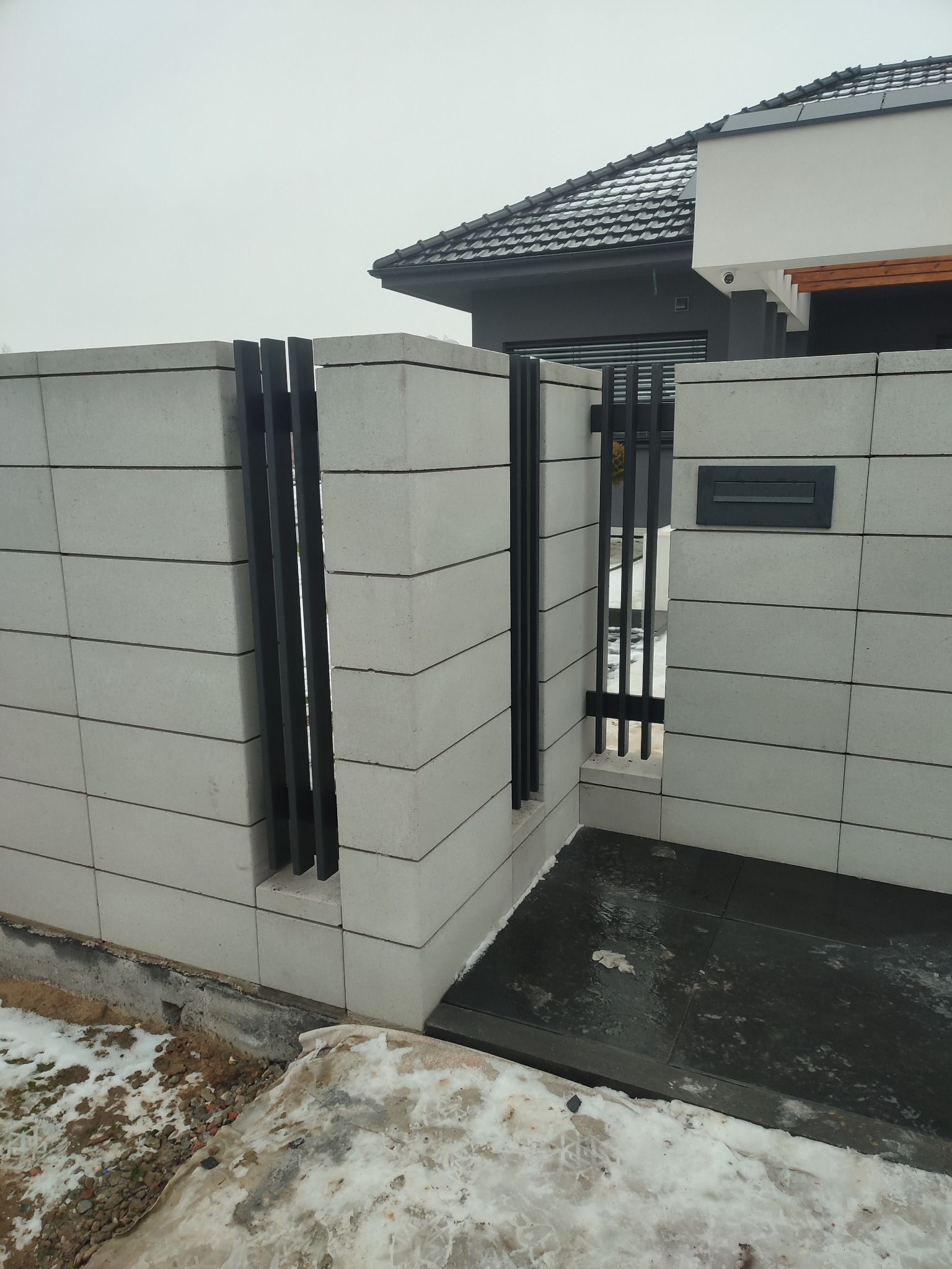Nowoczesne ogrodzenie z szarych betonowych bloków i czarnych, pionowych elementów metalowych, z widoczną skrzynką na listy i fragmentem domu w tle.