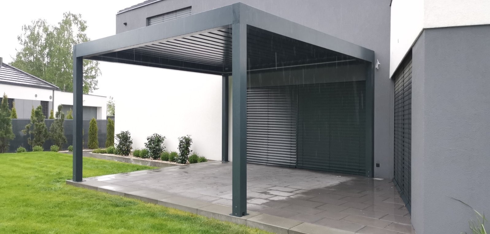 Nowoczesna pergola z metalową konstrukcją i lamelami na dachu, przylegająca do budynku z zamkniętymi żaluzjami fasadowymi w deszczowy dzień.