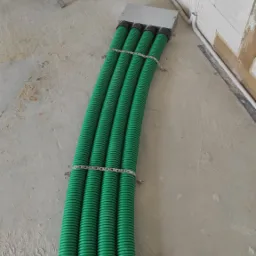 Cztery zielone, karbowane rury wentylacyjne prowadzone w podłodze, połączone metalowymi opaskami, w surowym stanie budynku z widocznymi pustakami i przewodami elektrycznymi.