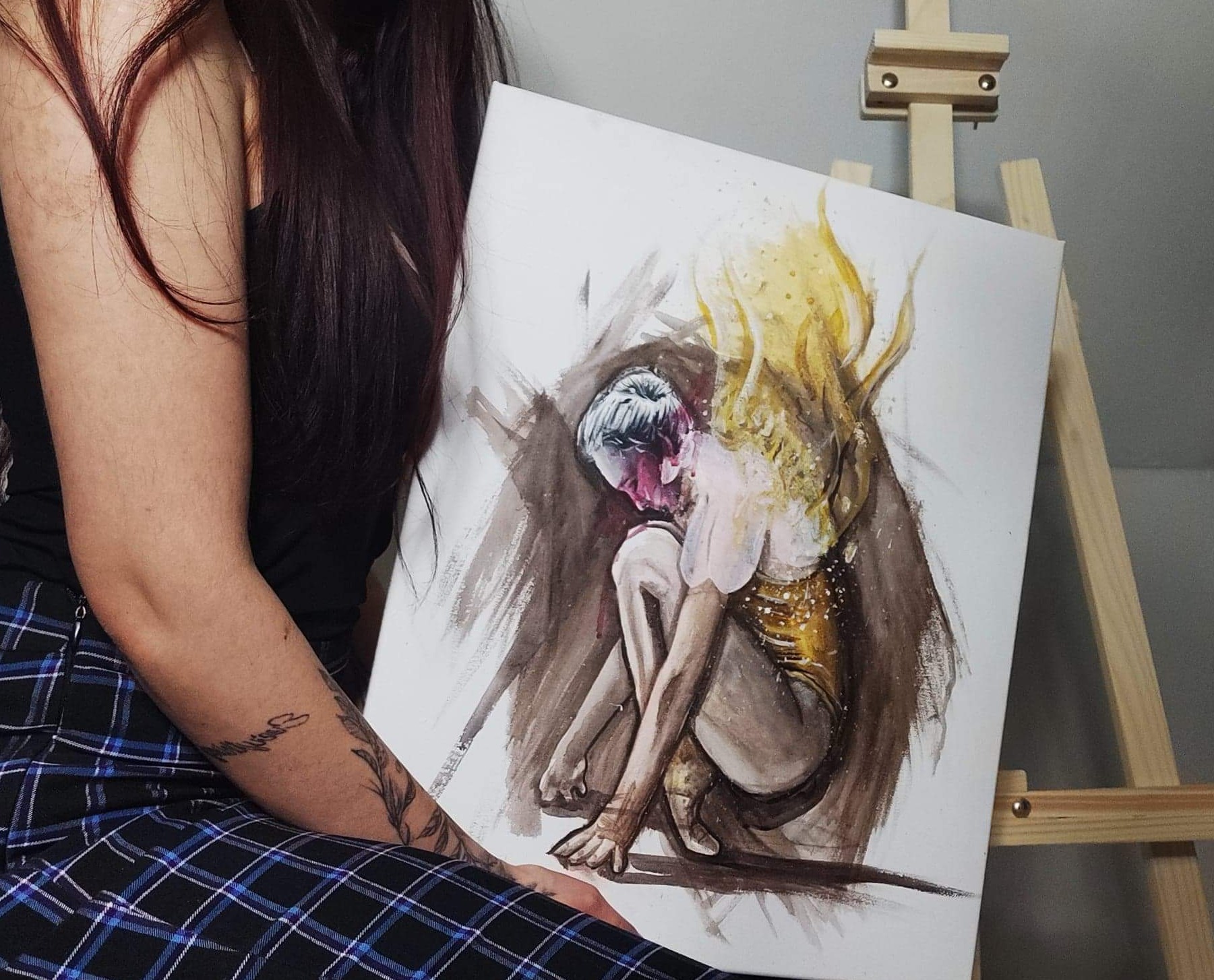 Kobieta trzyma akwarelowy portret skulonej postaci z jasnymi plamami koloru, ustawiony na drewnianej sztaludze; widoczny tatuaż na ramieniu kobiety.