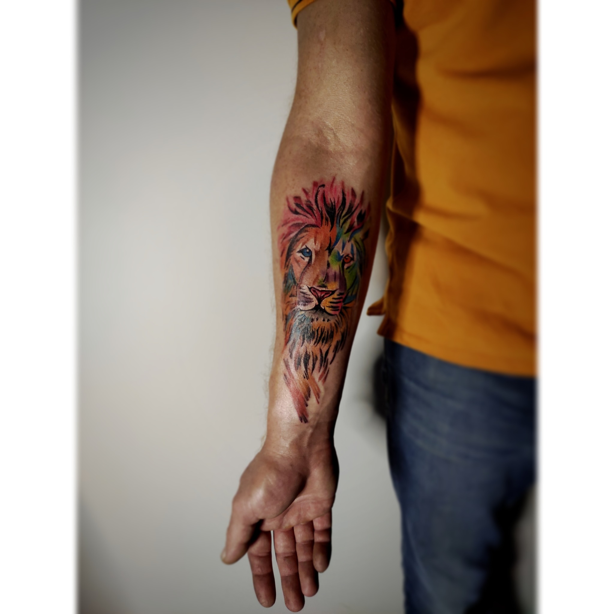 Ramię z kolorowym tatuażem lwa w stylu akwareli, widoczne żyły i zmarszczki na skórze, osoba ubrana w żółtą koszulkę i jeansy.