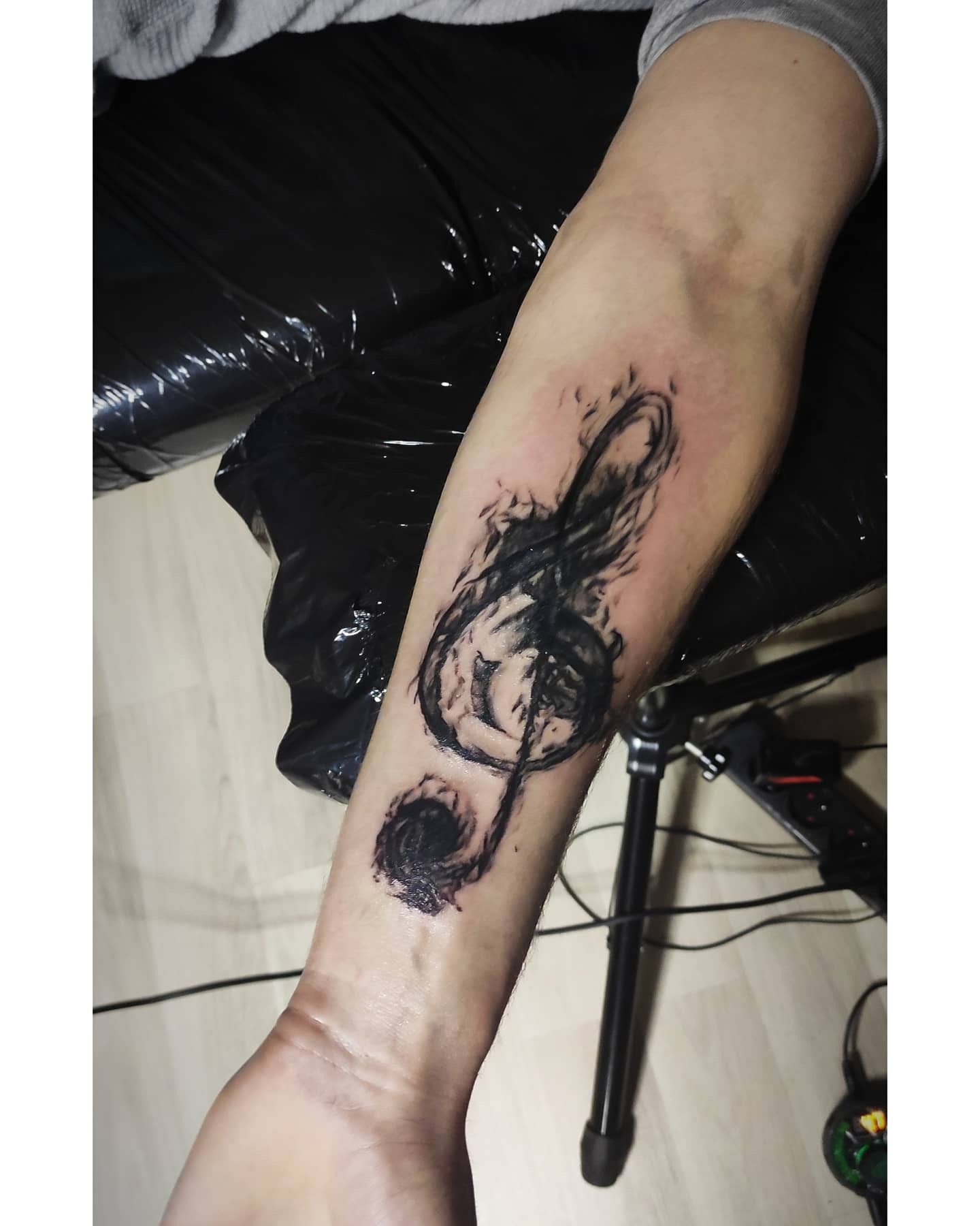 Tatuaż na przedramieniu przedstawiający klucz wiolinowy w stylu szkicu, z efektem rozmycia przypominającym dym, wykonany czarnym tuszem. Widoczny fragment statywu i czarnej folii w tle.