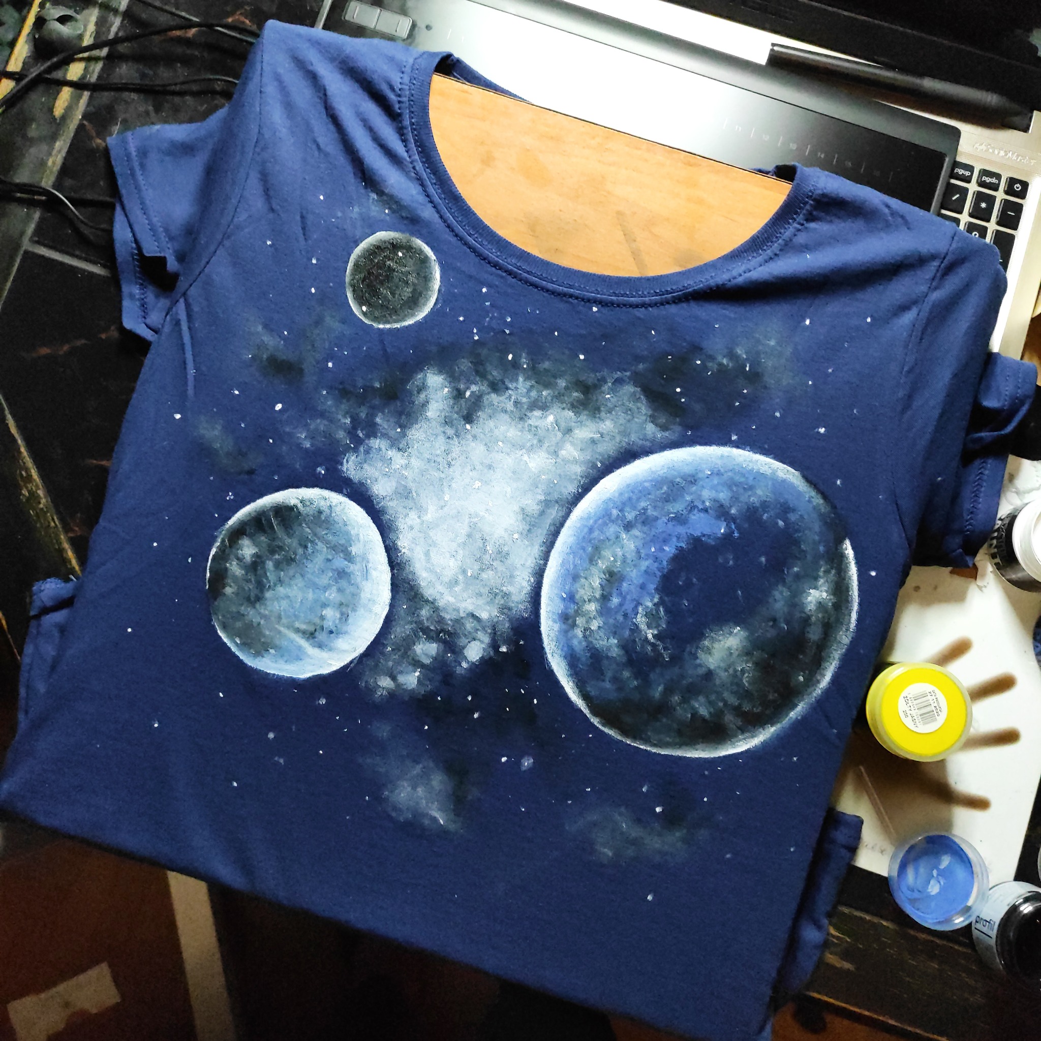 Granatowa koszulka z ręcznie malowanym motywem kosmosu: planety, gwiazdy i mgławice, leżąca obok otwartych słoiczków z farbami i tabletu graficznego.