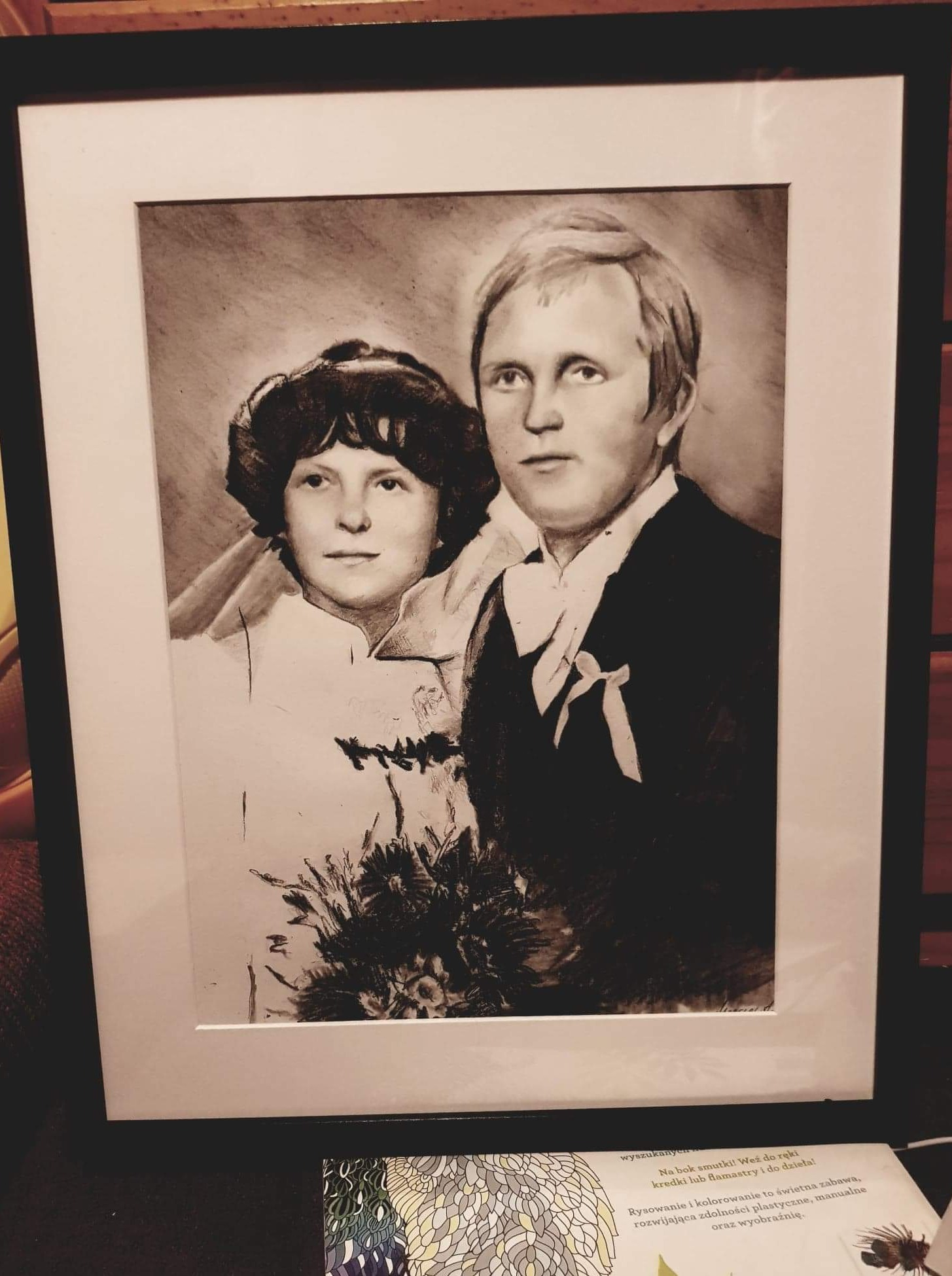 Portret ślubny pary młodej, wykonany w technice rysunku ołówkiem, oprawiony w czarną ramę, widoczny w ciepłym świetle wnętrza.