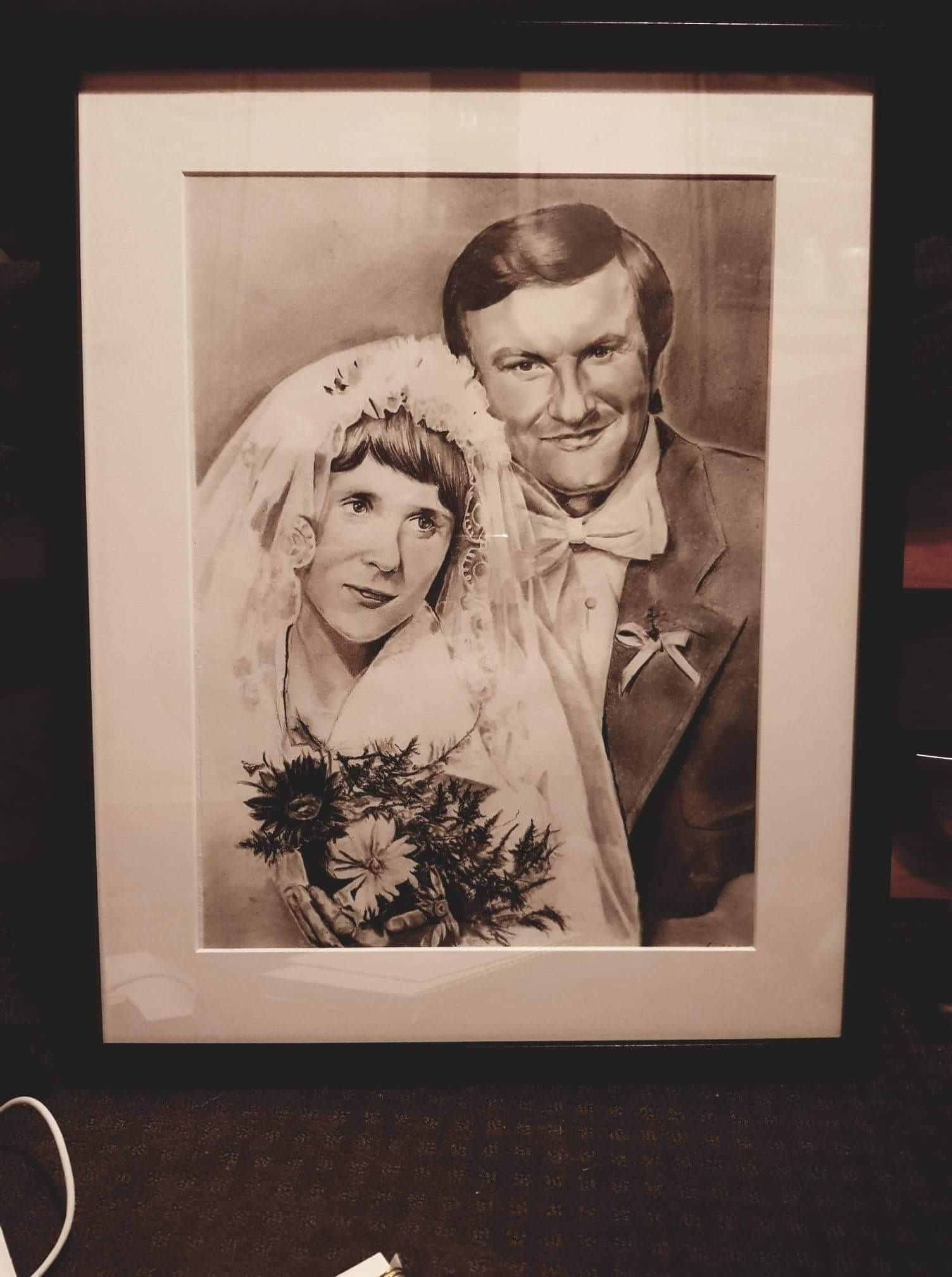 Oprawiony portret ślubny pary, wykonany w technice ołówkowej, przedstawia pannę młodą w welonie i z bukietem oraz pana młodego w garniturze z muchą.