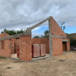 Wznoszenie ścian parterowego domu z czerwonej cegły ceramicznej, widoczne pakiety cegieł na paletach wewnątrz budynku, szary betonowy szkielet konstrukcyjny, na zewnątrz piaszczysta ziemia...