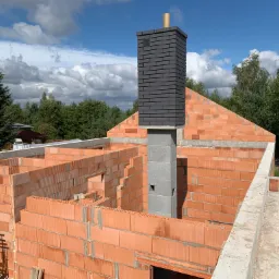 Wznoszenie ścian z czerwonej cegły ceramicznej domu w trakcie budowy, widoczny komin z szarej cegły klinkierowej i niebieskie niebo z chmurami w tle.