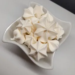 Gwiazdkowe bezy w białej, kwadratowej miseczce o falistych brzegach, na jasnym tle, z widoczną delikatną strukturą cukierniczą.