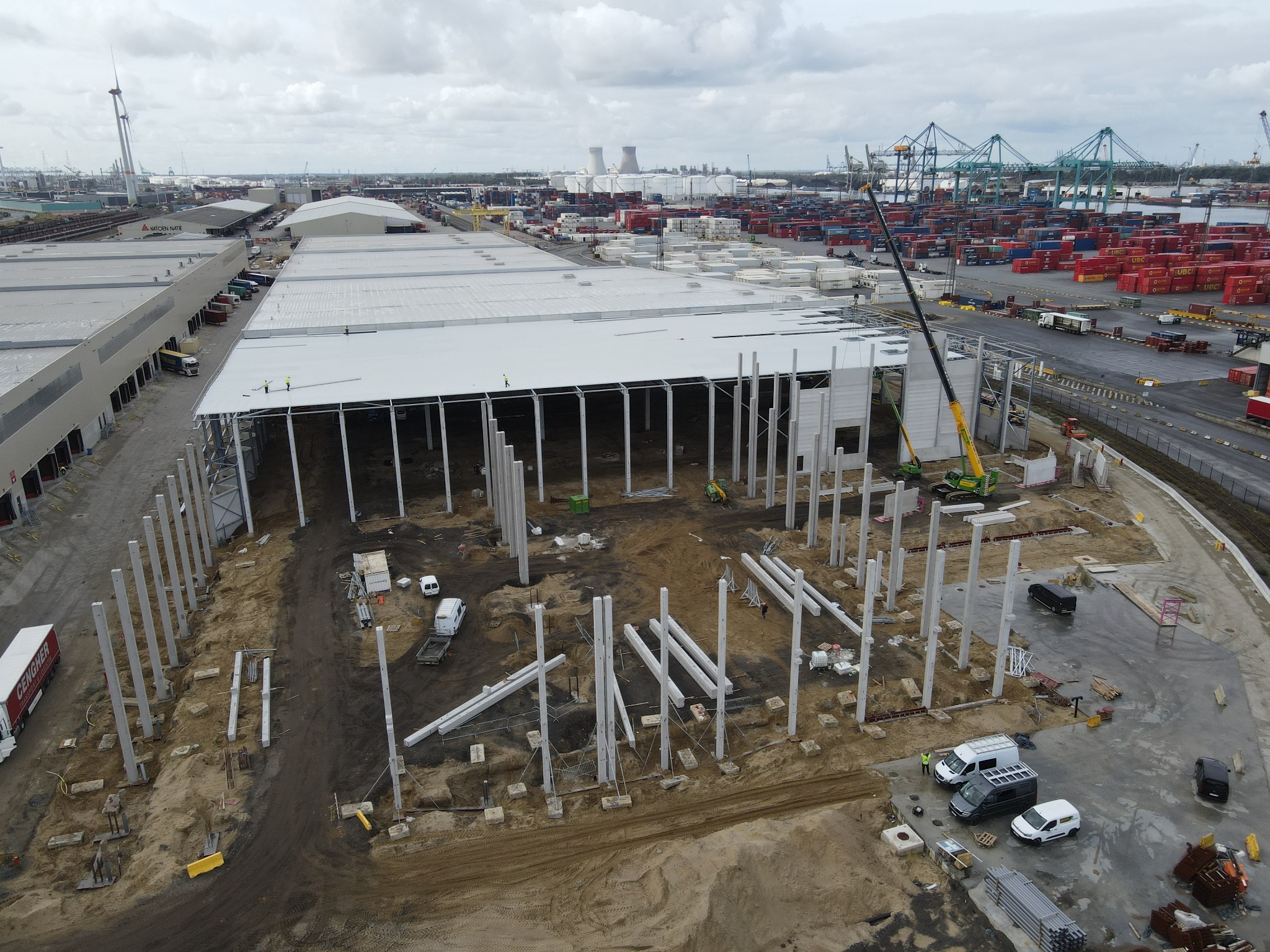 Szeroki kadr z lotu ptaka na plac budowy hali magazynowej w porcie, z widocznymi betonowymi słupami konstrukcyjnymi, częściowo zamontowanym dachem z płyt warstwowych, dźwigiem, ciężarówkami...