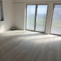 Jasne panele podłogowe ułożone w nowym, pustym pomieszczeniu z dużymi oknami zabezpieczonymi folią, widoczny sprzęt glazurniczy w rogu.