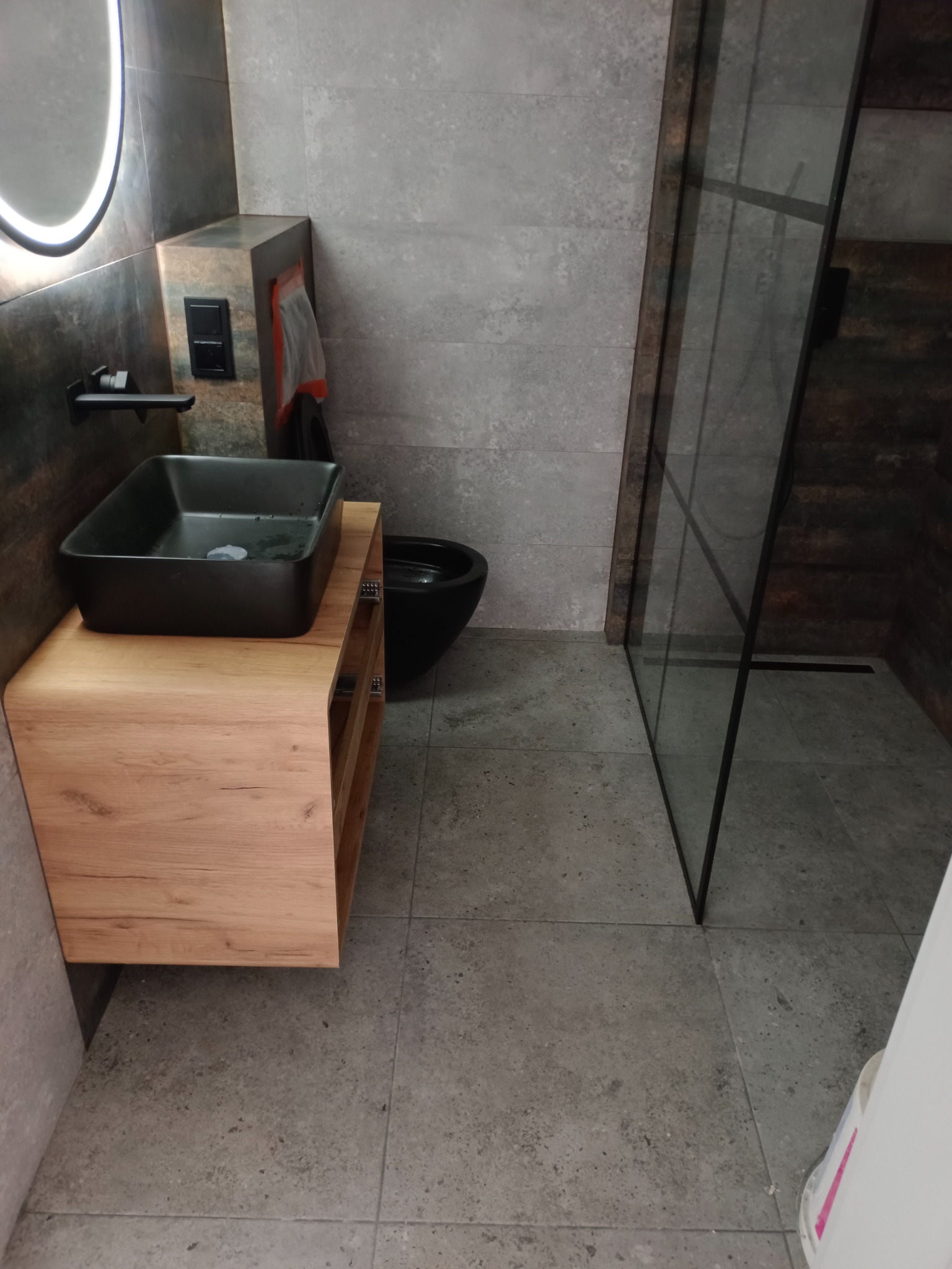 Nowoczesna łazienka z czarną umywalką nablatową na drewnianej szafce, szarymi płytkami i kabiną prysznicową z czarnymi ramami.