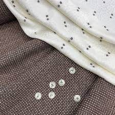 Biała tkanina z drobnym wzorem, ułożona na brązowej tkaninie w kratkę, z rozsypanymi białymi guzikami z czterema dziurkami.