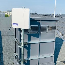 Instal-SAT Damian Moskal - Antena operatora Play zamontowana na dachu budynku, przymocowana do szarej konstrukcji z wentylacją, widok na dach pokryty papą i błękitne niebo.