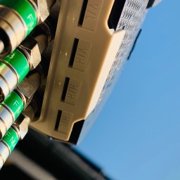 Instal-SAT Damian Moskal - Złącza antenowe F-MA z zielonymi pierścieniami, podłączone do białego rozdzielacza antenowego na tle błękitnego nieba.