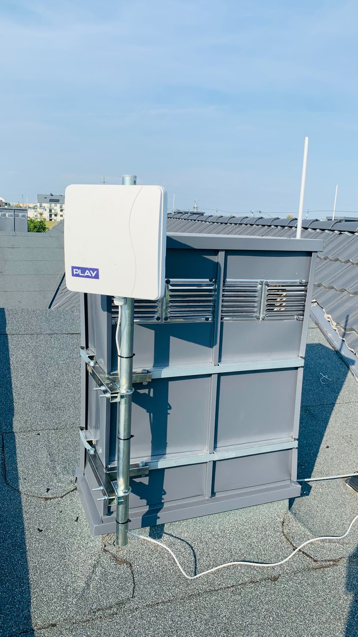 Antena operatora Play zamontowana na dachu budynku, przymocowana do szarej konstrukcji z wentylacją, widok na dach pokryty papą i błękitne niebo.