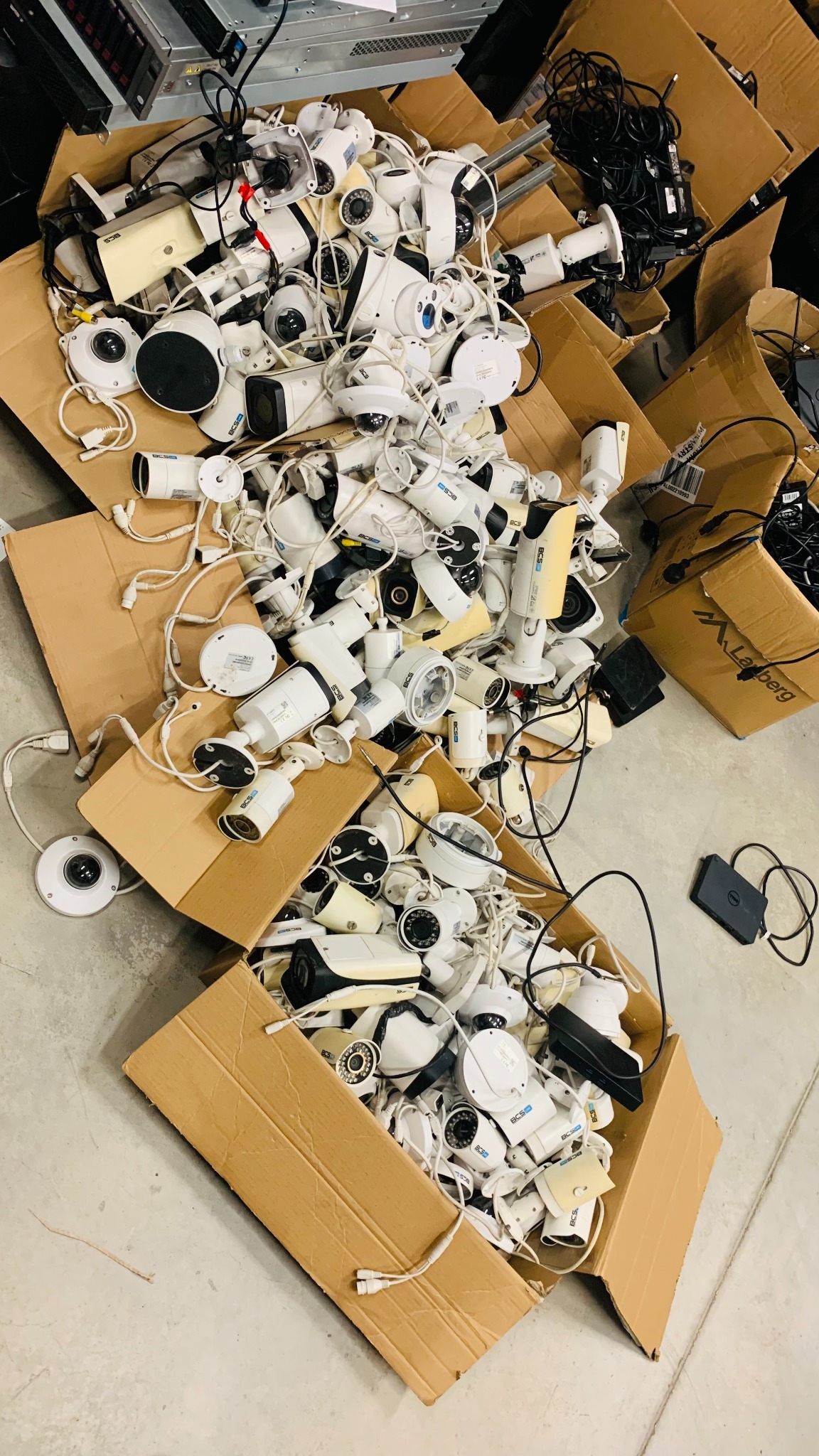 Kartonowe pudła wypełnione białymi kamerami przemysłowymi różnych typów i rozmiarów, wraz z kablami i akcesoriami, widok z góry.