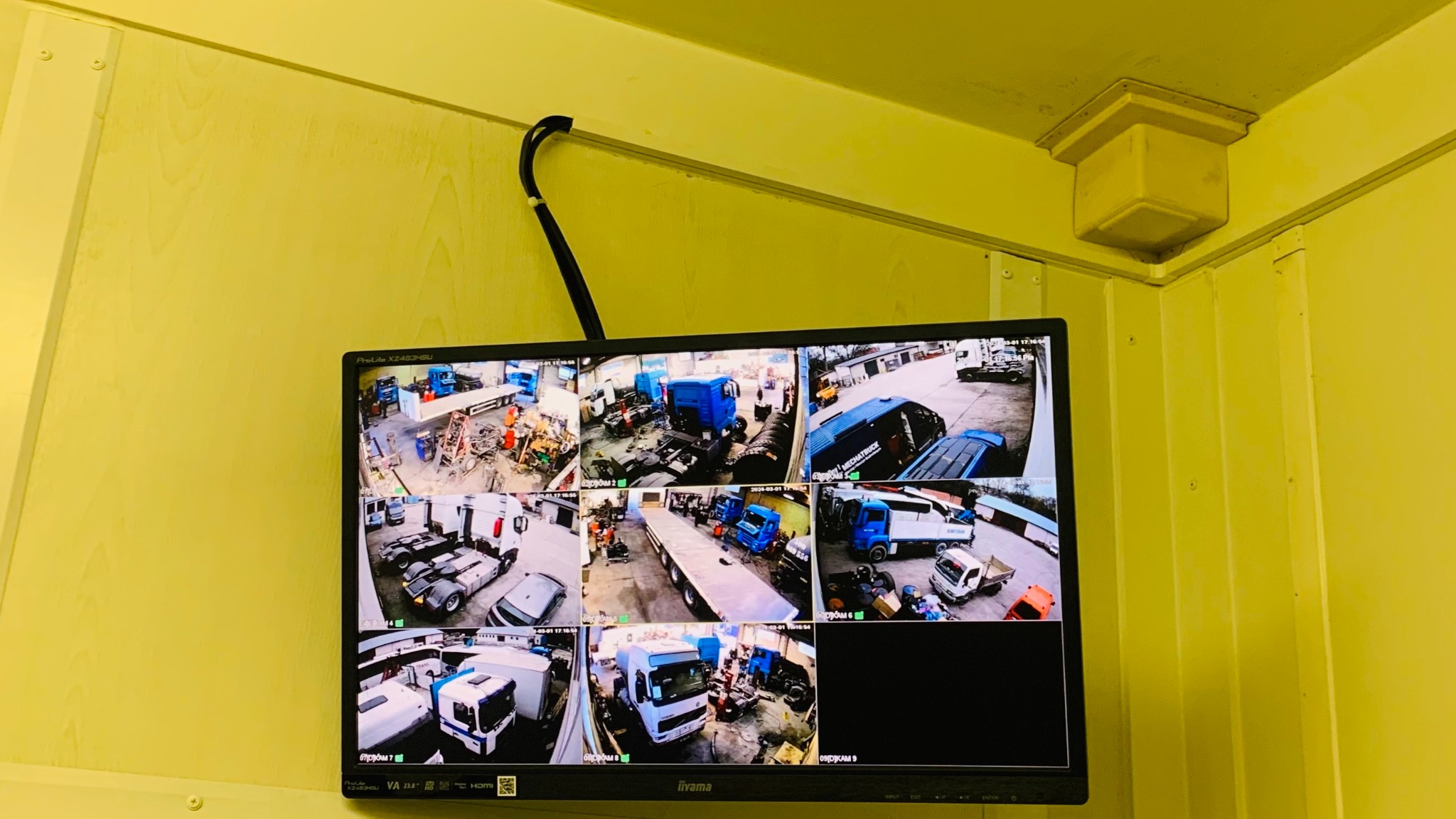 Ekran monitora CCTV zamontowanego na żółtej ścianie, wyświetlający podgląd z dziewięciu kamer monitoringu, ukazujących prace serwisowe przy ciężarówkach i naczepach w warsztacie.