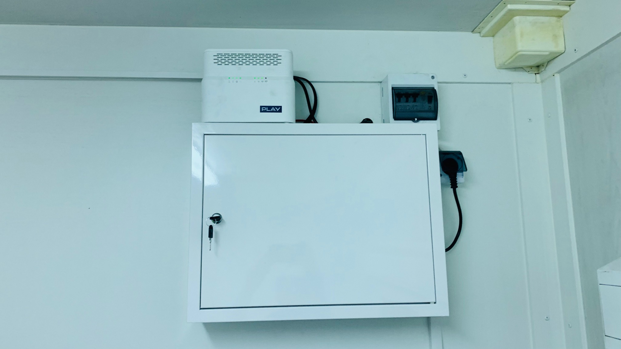 Biała szafka zamykana na klucz, router Play z diodami LED, skrzynka bezpiecznikowa i gniazdko elektryczne zamontowane na białej ścianie.