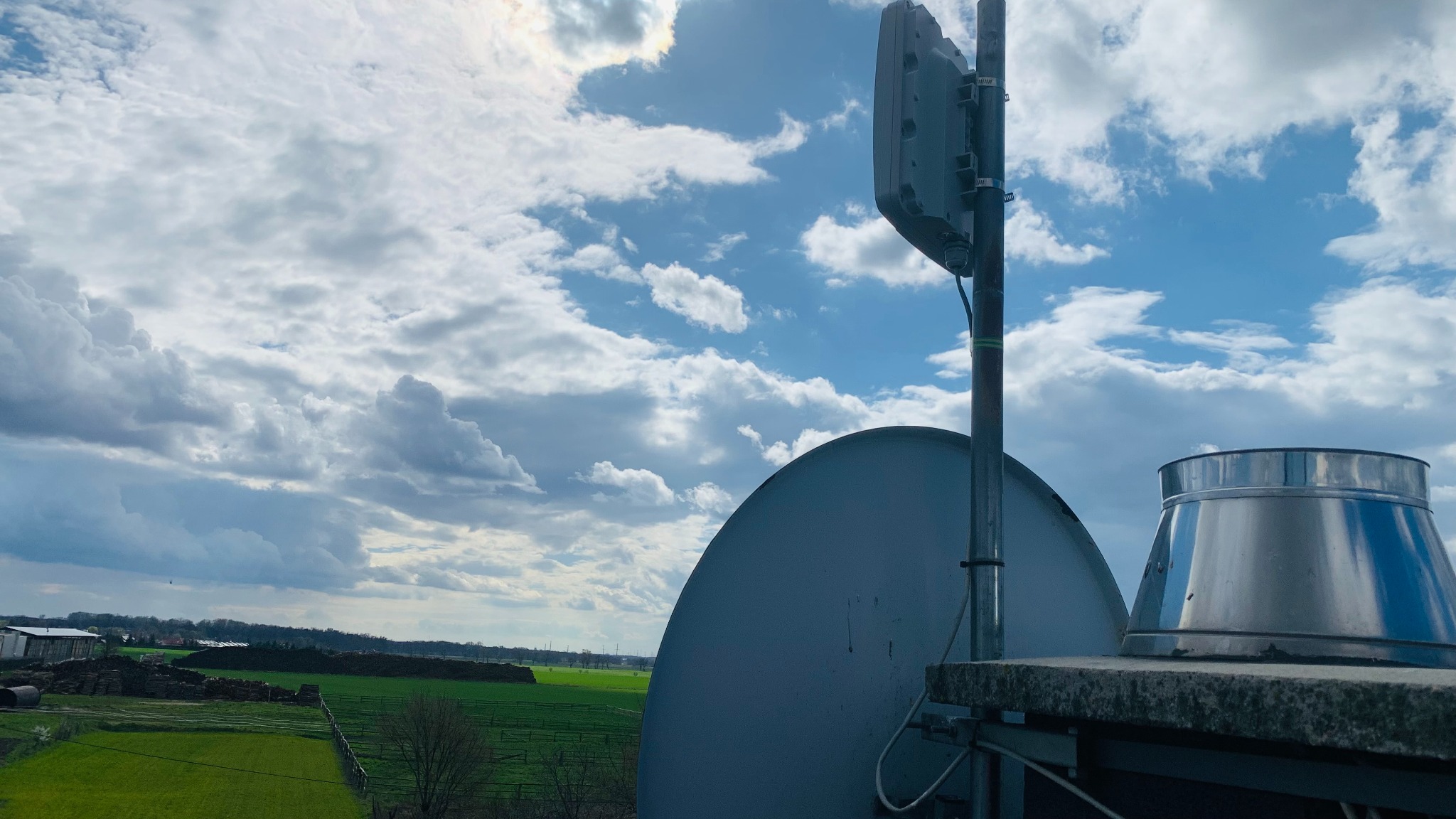 Antena satelitarna i dodatkowa instalacja na dachu budynku we Wrocławiu, z widokiem na pola i zachmurzone niebo.