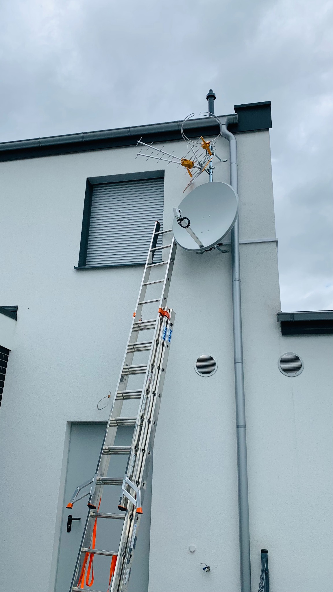 Antena satelitarna i antena naziemna zamontowane na elewacji budynku, z przystawioną aluminiową drabiną Krause z pomarańczowymi zabezpieczeniami.