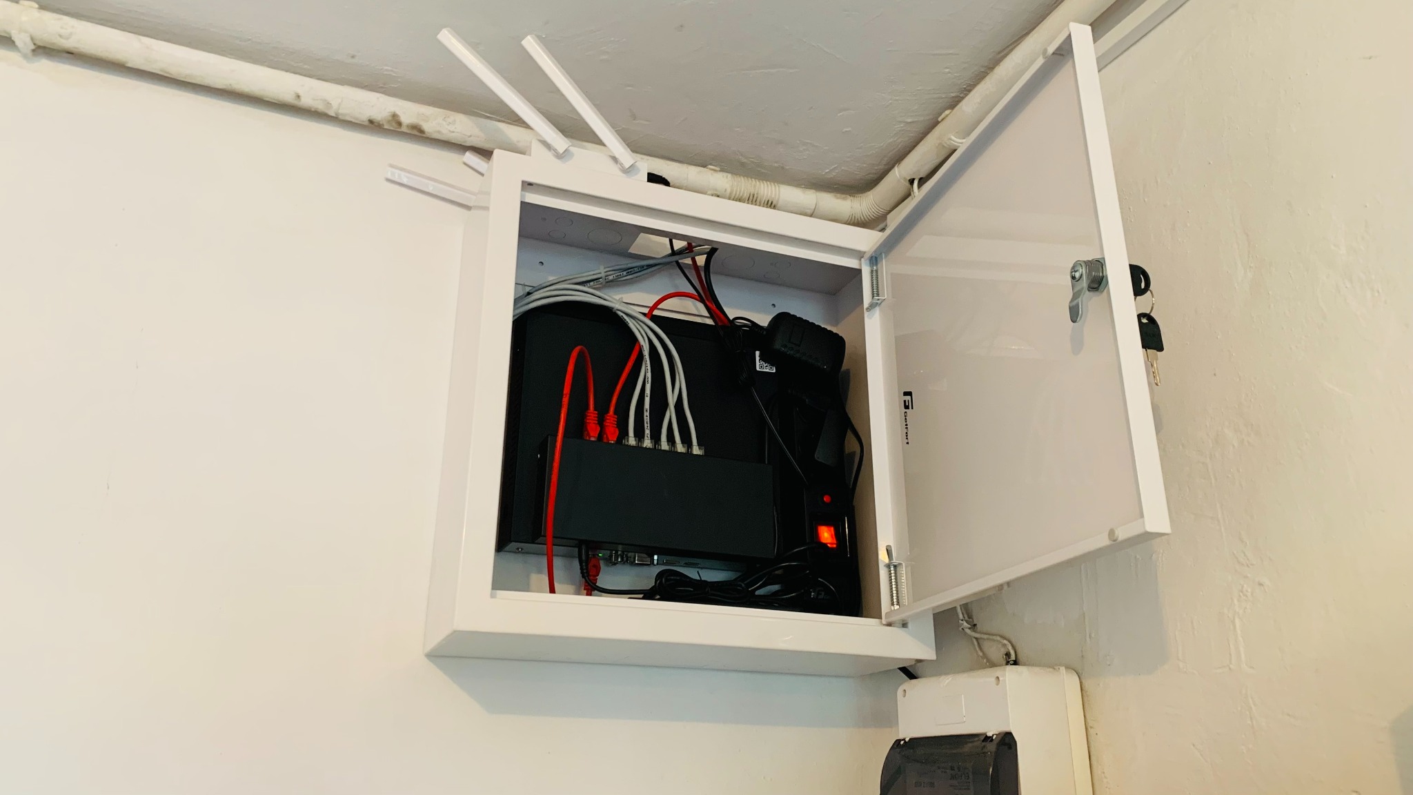 Szafka telekomunikacyjna z otwartymi drzwiczkami, widoczny switch sieciowy, kable Ethernet w kolorach czerwonym i białym, antena Wi-Fi na górze, klucze wiszące na zamku, instalacja wewnątrz budynku.