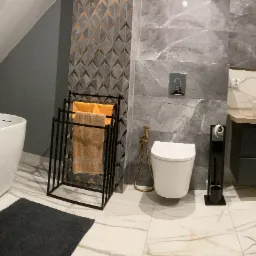 Nowoczesna łazienka na poddaszu z białą wolnostojącą wanną, marmurowymi płytkami, złotą armaturą i czarnymi dodatkami, oświetlona światłem z okna dachowego.