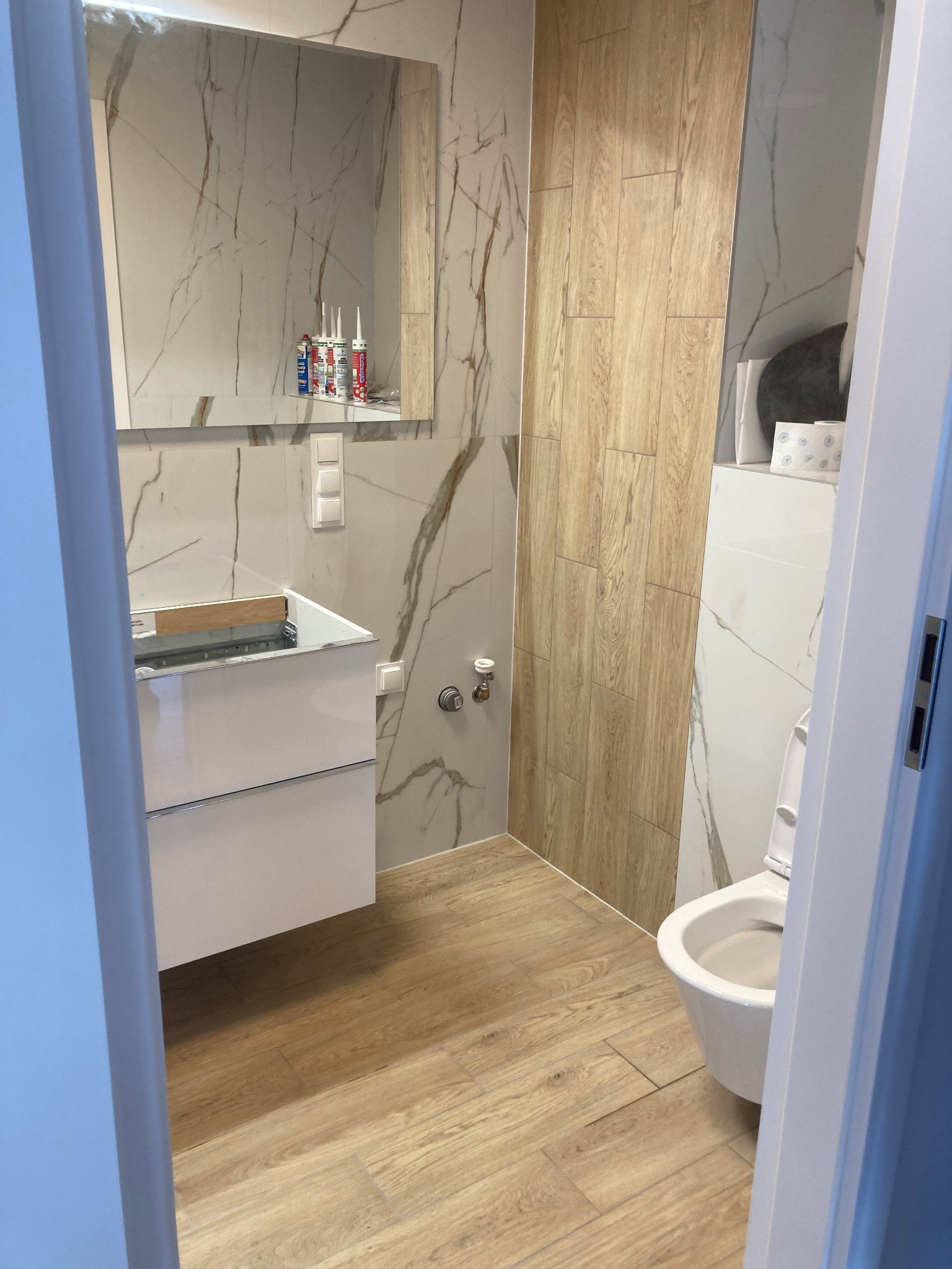 Wnętrze łazienki w trakcie remontu, widoczna biała umywalka z szafką, toaleta, płytki imitujące drewno i marmur oraz silikony na półce pod lustrem.