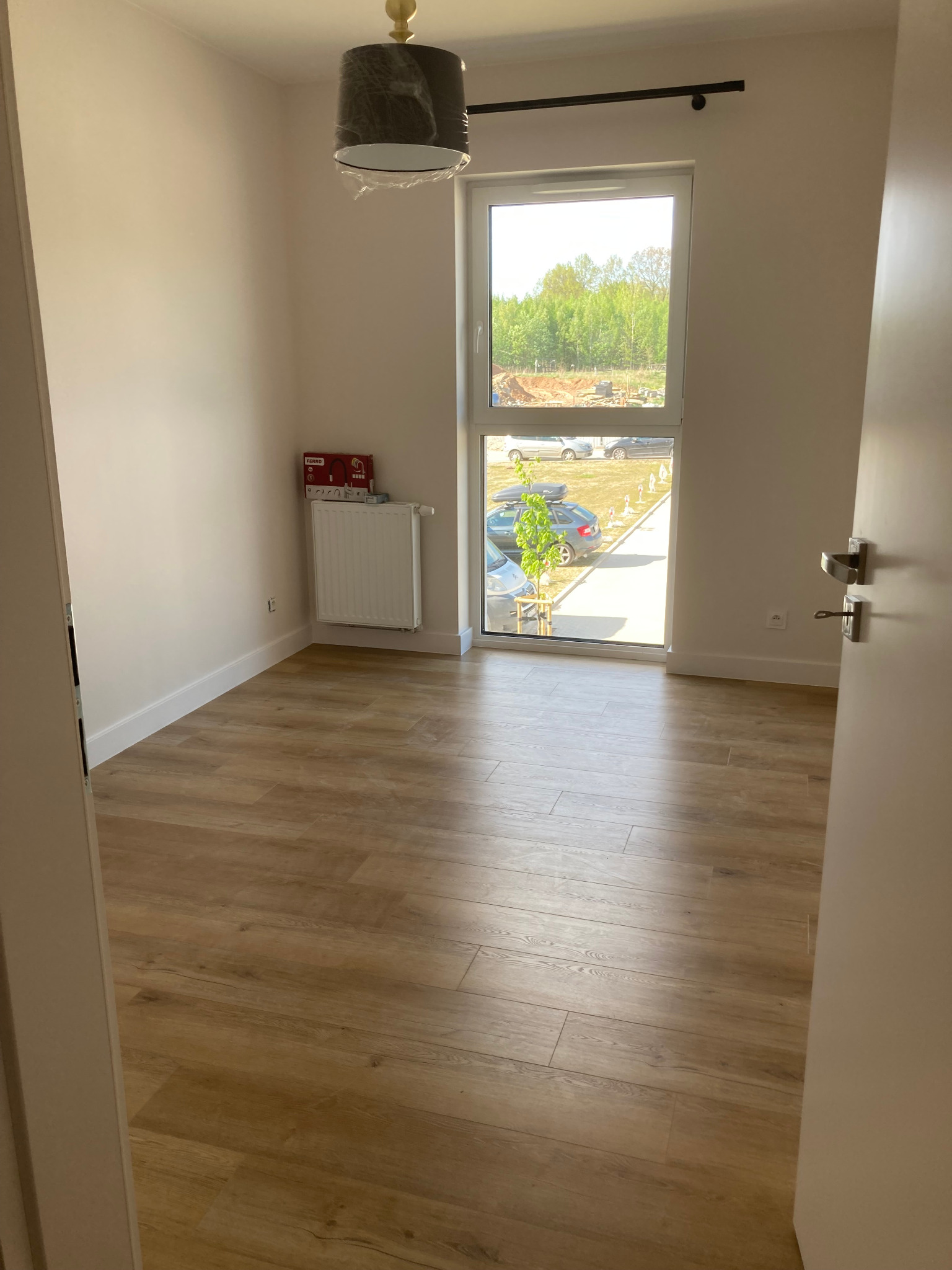 Pusty pokój po remoncie z panelami podłogowymi imitującymi drewno, białymi ścianami, grzejnikiem, oknem z widokiem na parking i lampą wiszącą w folii ochronnej.