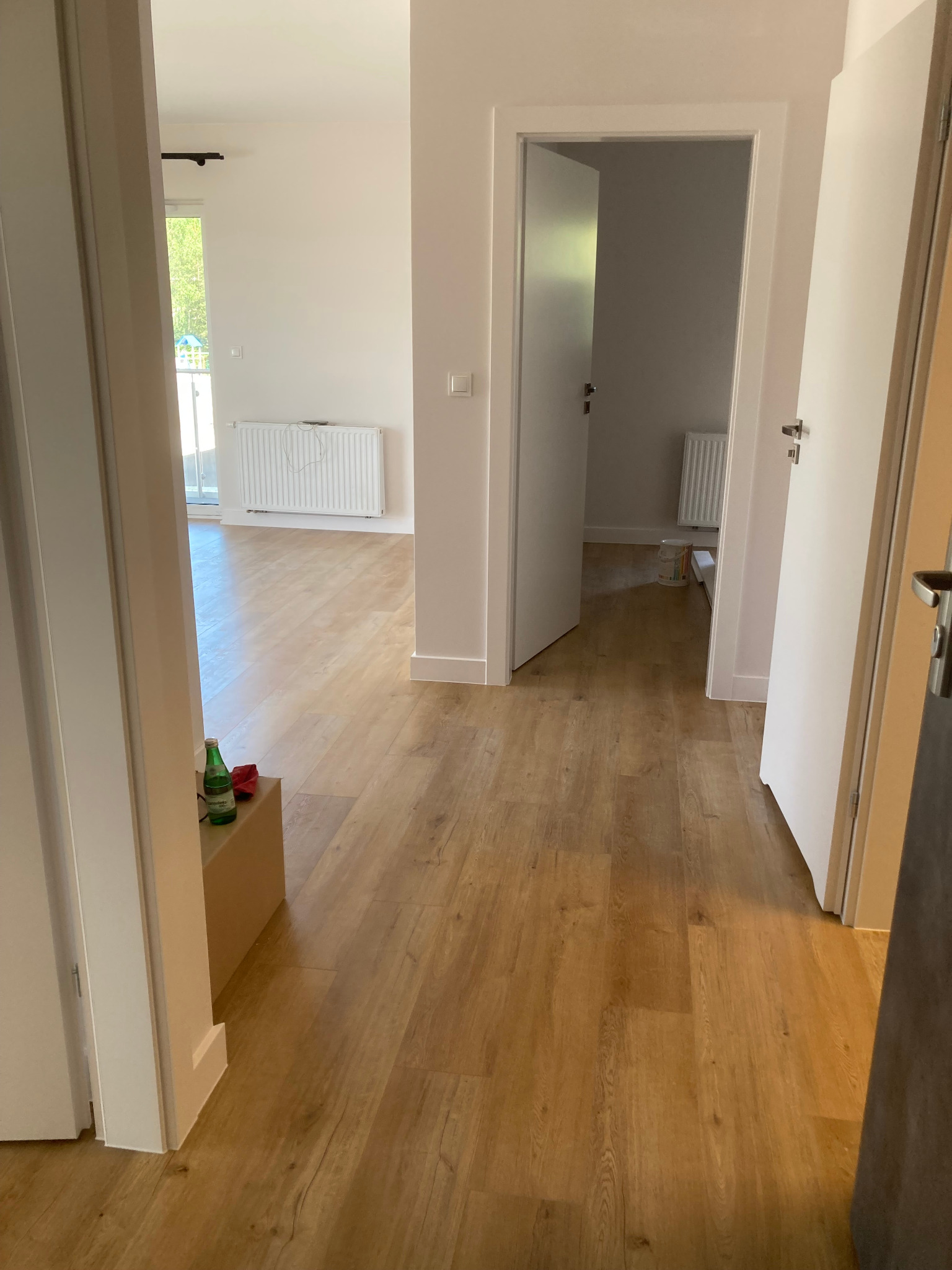 Widok z perspektywy wnętrza mieszkania na świeżo ułożone panele podłogowe, z widocznymi drzwiami wejściowymi, butelką wody mineralnej przy ścianie i grzejnikiem w tle, sugerujący etap końcowy prac...