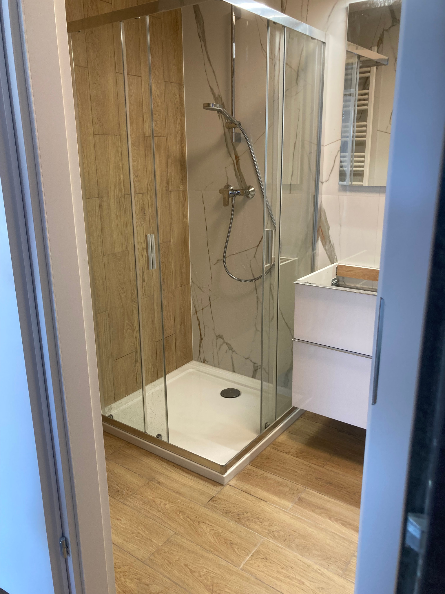 Nowoczesna kabina prysznicowa z drzwiami przesuwnymi i brodzikiem, widok zza uchylonych drzwi, płytki imitujące drewno na ścianie, marmurowe wykończenie ściany za kabiną, biała szafka pod umywalkę...