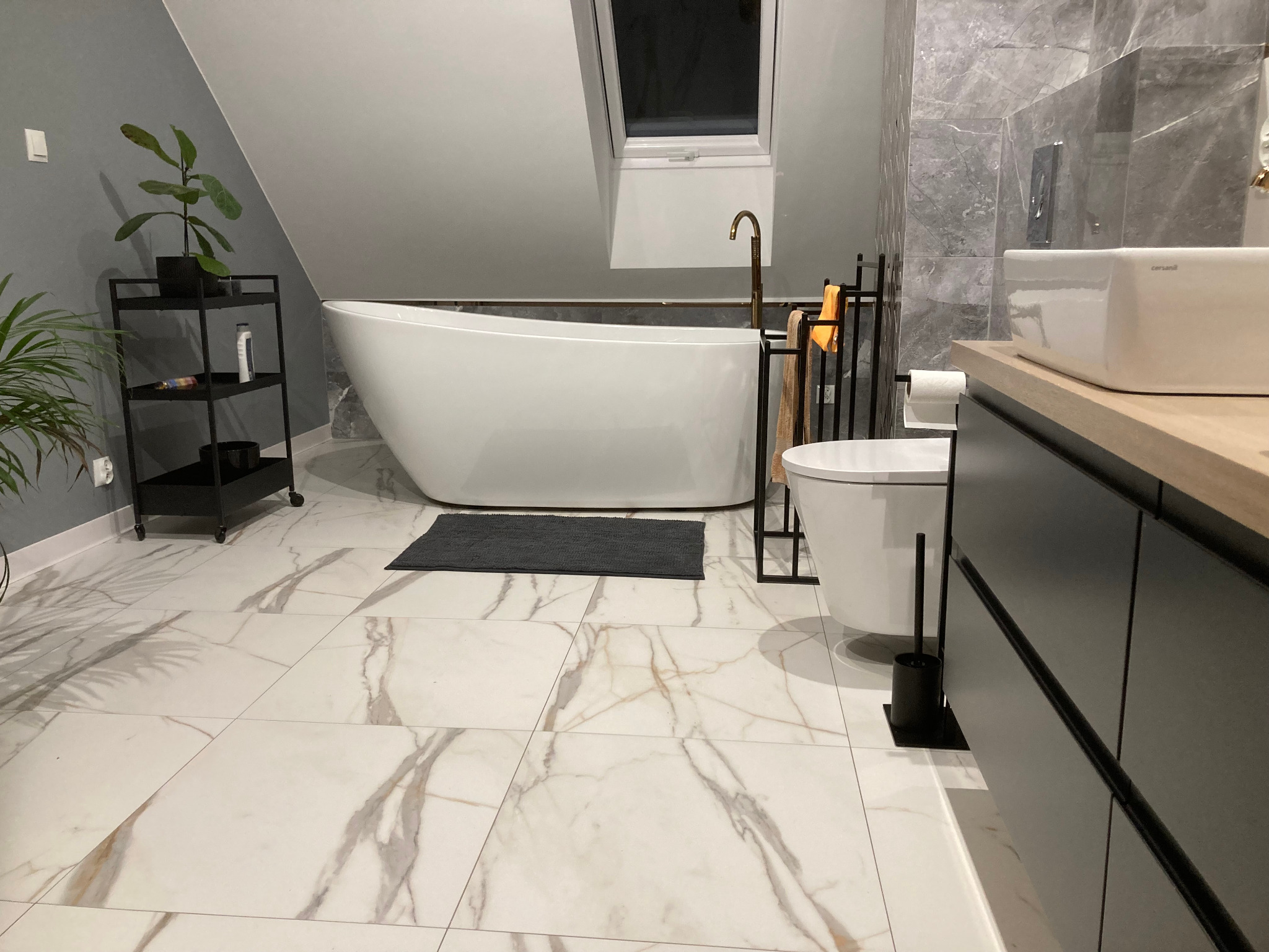 Nowoczesna łazienka na poddaszu z wolnostojącą wanną, marmurową podłogą i szarą armaturą, oświetlona przez okno dachowe.