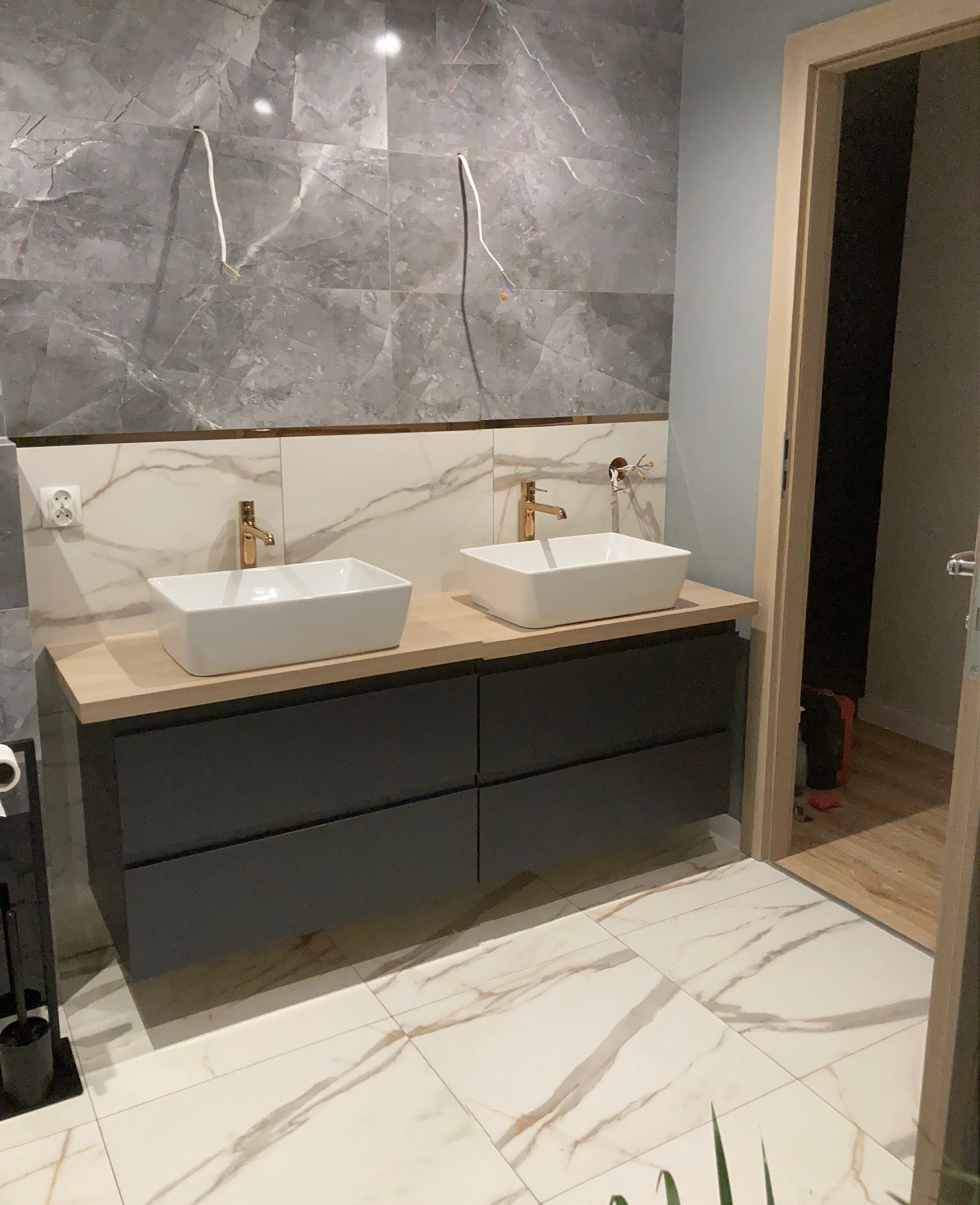 Nowoczesna łazienka w trakcie wykończenia: dwie białe umywalki nablatowe z złotymi bateriami na szarej szafce, marmurowe płytki na ścianie z wystającymi kablami, podłoga w jasne płytki z marmurowym...
