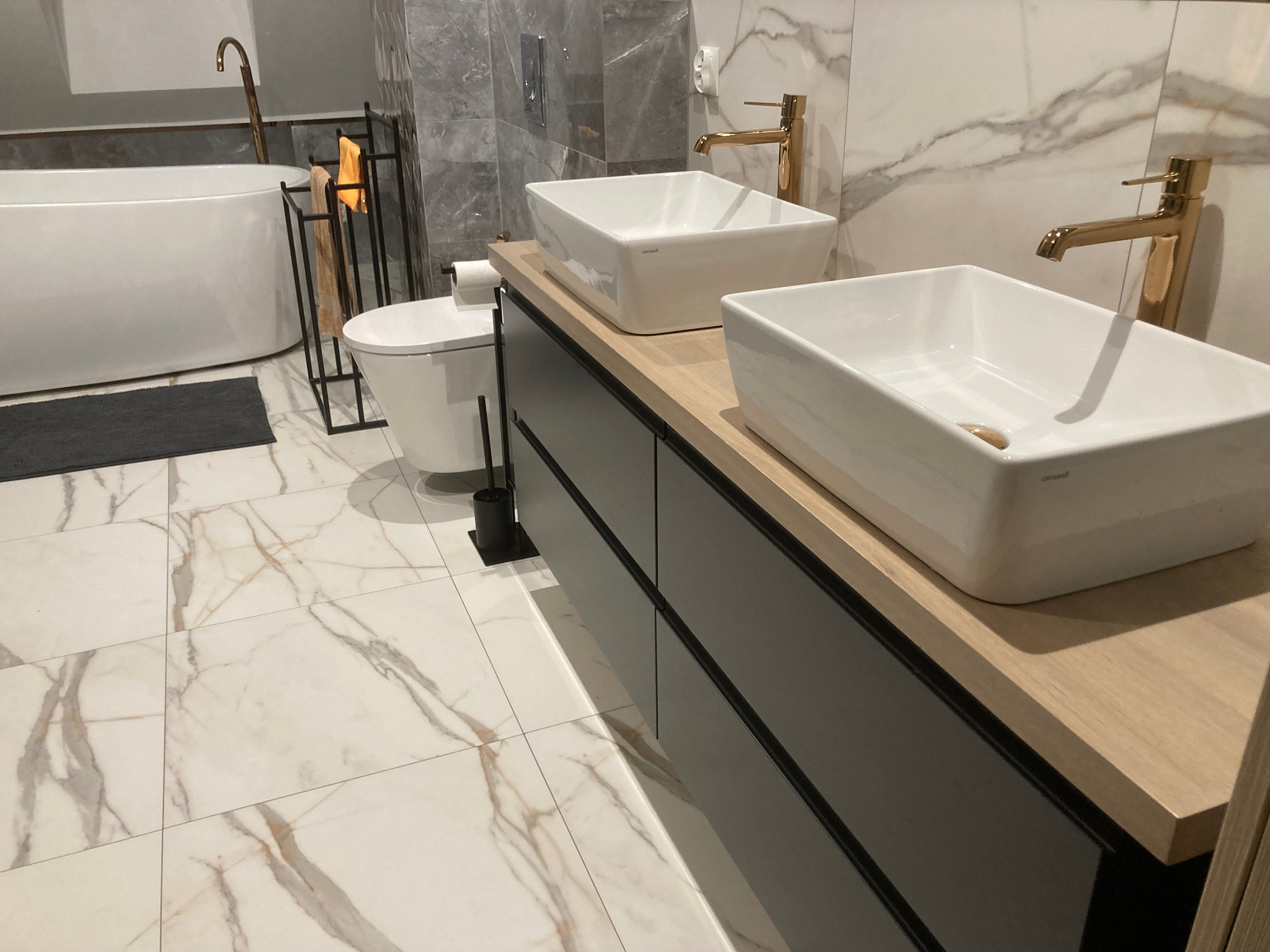 Nowoczesna łazienka w Kudowa-Zdrój z marmurowymi płytkami, dwiema białymi umywalkami nablatowymi z bateriami w kolorze złotym, wiszącą toaletą i wolnostojącą wanną.