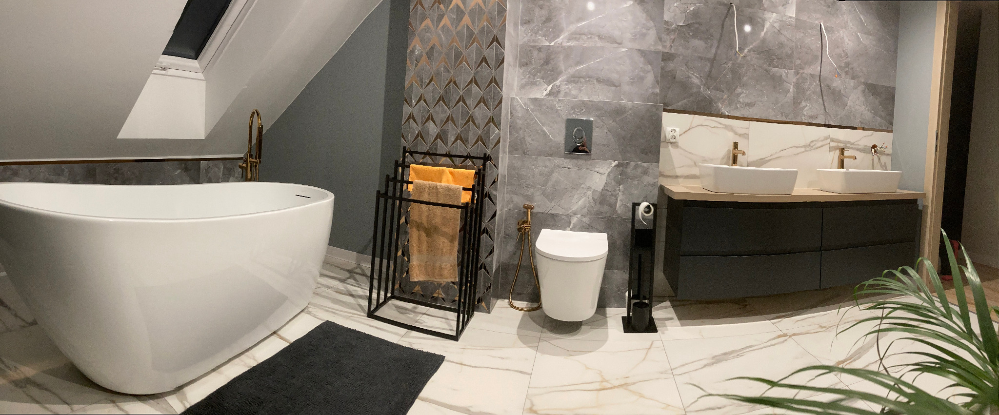 Nowoczesna łazienka na poddaszu z białą wolnostojącą wanną, marmurowymi płytkami, złotą armaturą i czarnymi dodatkami, oświetlona światłem z okna dachowego.