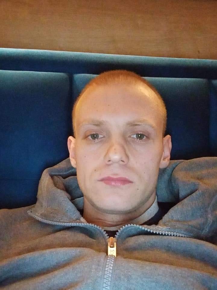 Portret mężczyzny z krótkimi, rudymi włosami, ubranego w szarą bluzę z kapturem na tle niebieskiej tapicerki.