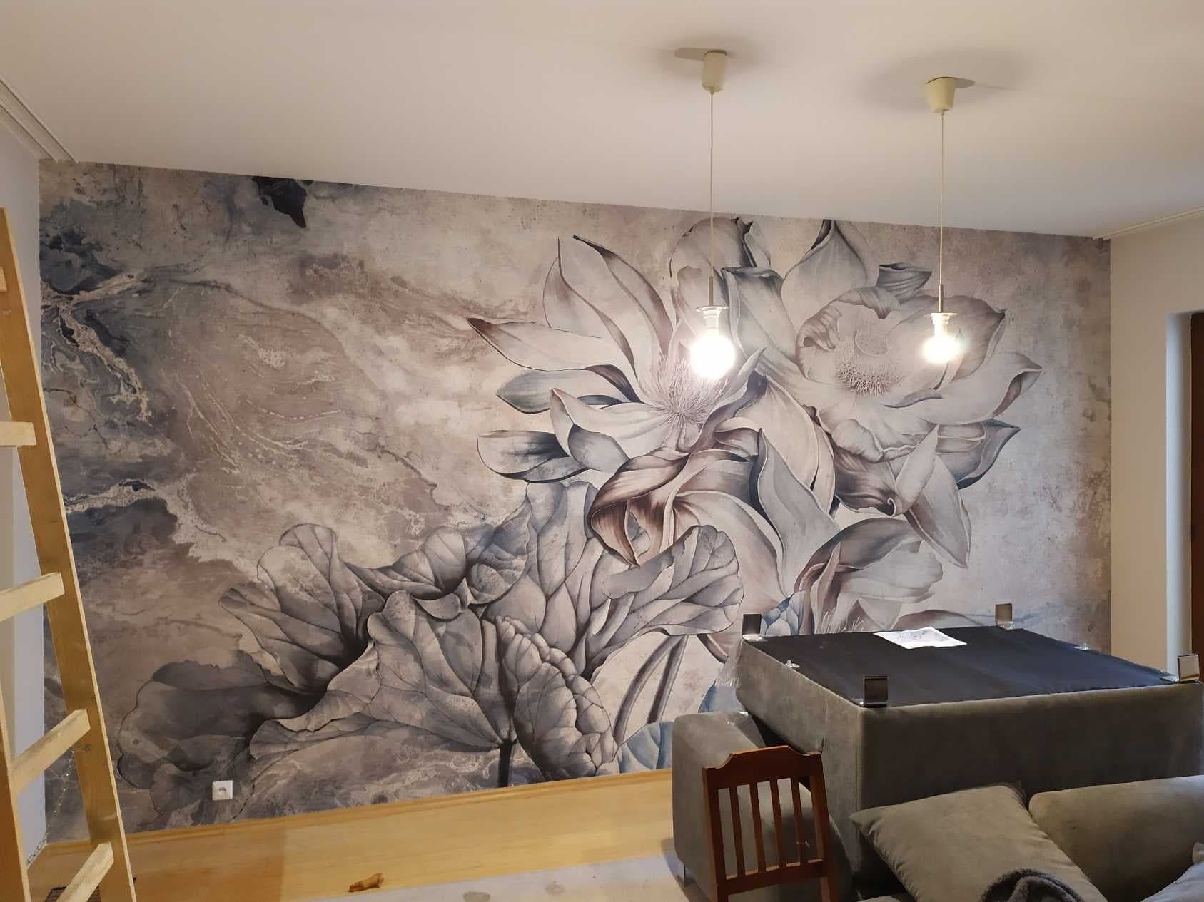 Ściana z fototapetą w odcieniach szarości z motywem kwiatowym, dwie wiszące lampy z żarówkami, drewniana drabina oparta o ścianę, fragment sofy z poduszkami.