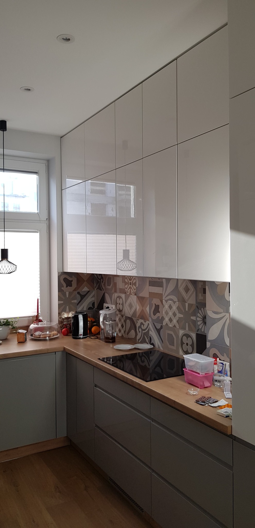 Nowoczesna kuchnia z szarymi szafkami na wysoki połysk, drewnianym blatem i płytkami w stylu patchwork za płytą indukcyjną. Na blacie czajnik, owoce i pojemniki plastikowe.