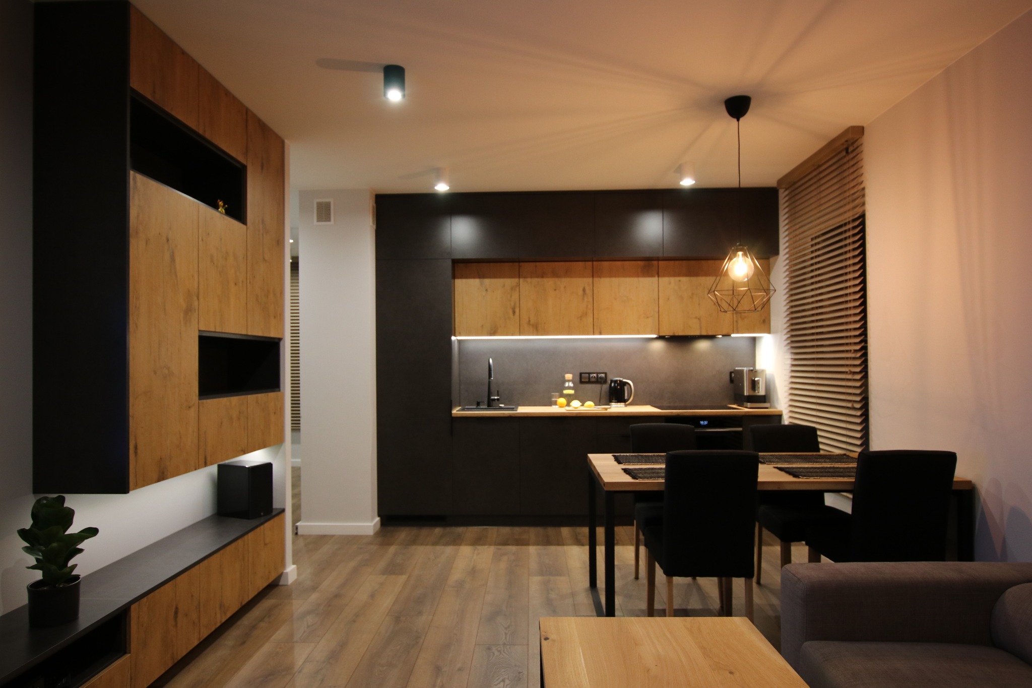 Nowoczesne wnętrze mieszkania: drewniana zabudowa ściany z czarnymi akcentami, otwarta kuchnia z jadalnią, geometryczna lampa wisząca, drewniana podłoga.
