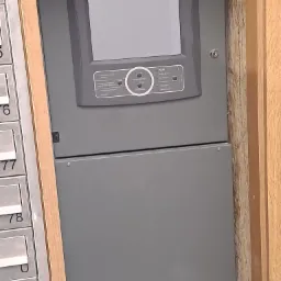 Szara centrala alarmowa Polon-Alfa zamontowana na ścianie, widoczna obok skrzynki pocztowej z numerami, z wyświetlaczem i przyciskami funkcyjnymi.