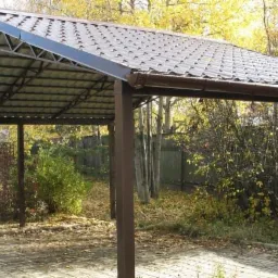 Zadaszenie tarasowe z metalową konstrukcją i brązową dachówką, widoczne z zewnątrz na tle jesiennej roślinności.