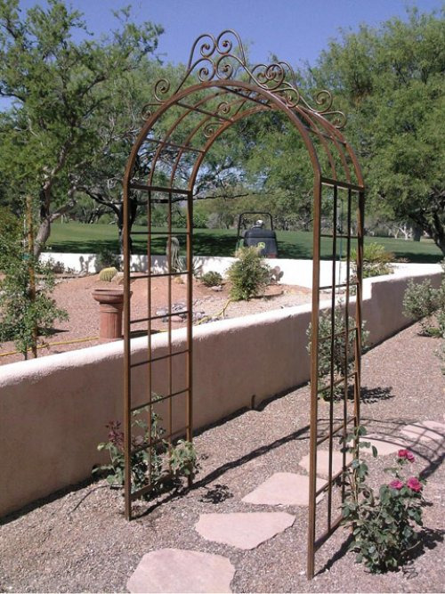 Metalowa pergola ogrodowa w rustykalnym stylu z dekoracyjnymi zawijasami, otoczona kamienną ścieżką i roślinnością, w tle trawnik i drzewa.